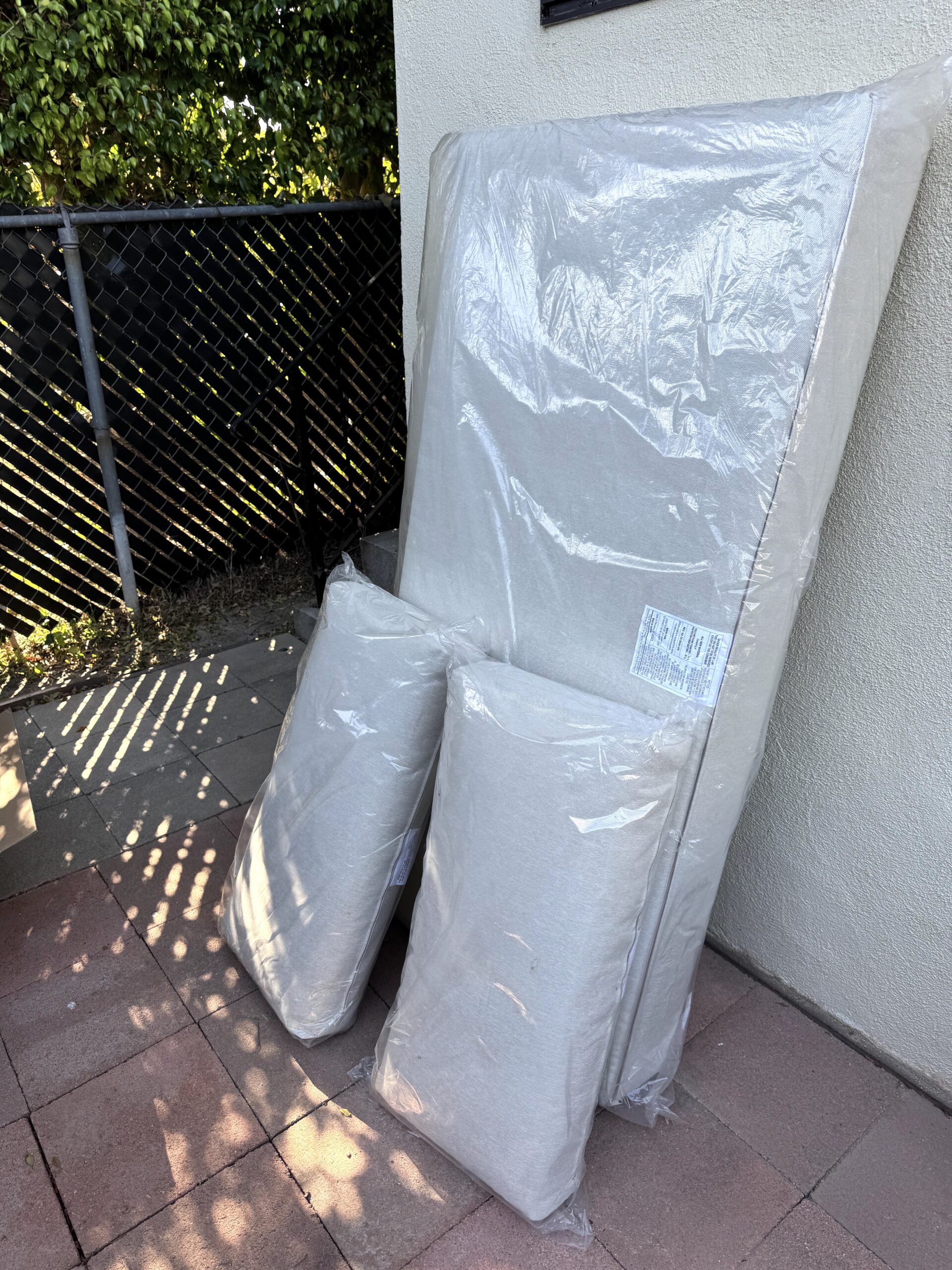 Two large cushions and two smaller cushions, all wrapped in clear plastic, are propped against an exterior wall on a tiled patio near a chain-link fence.