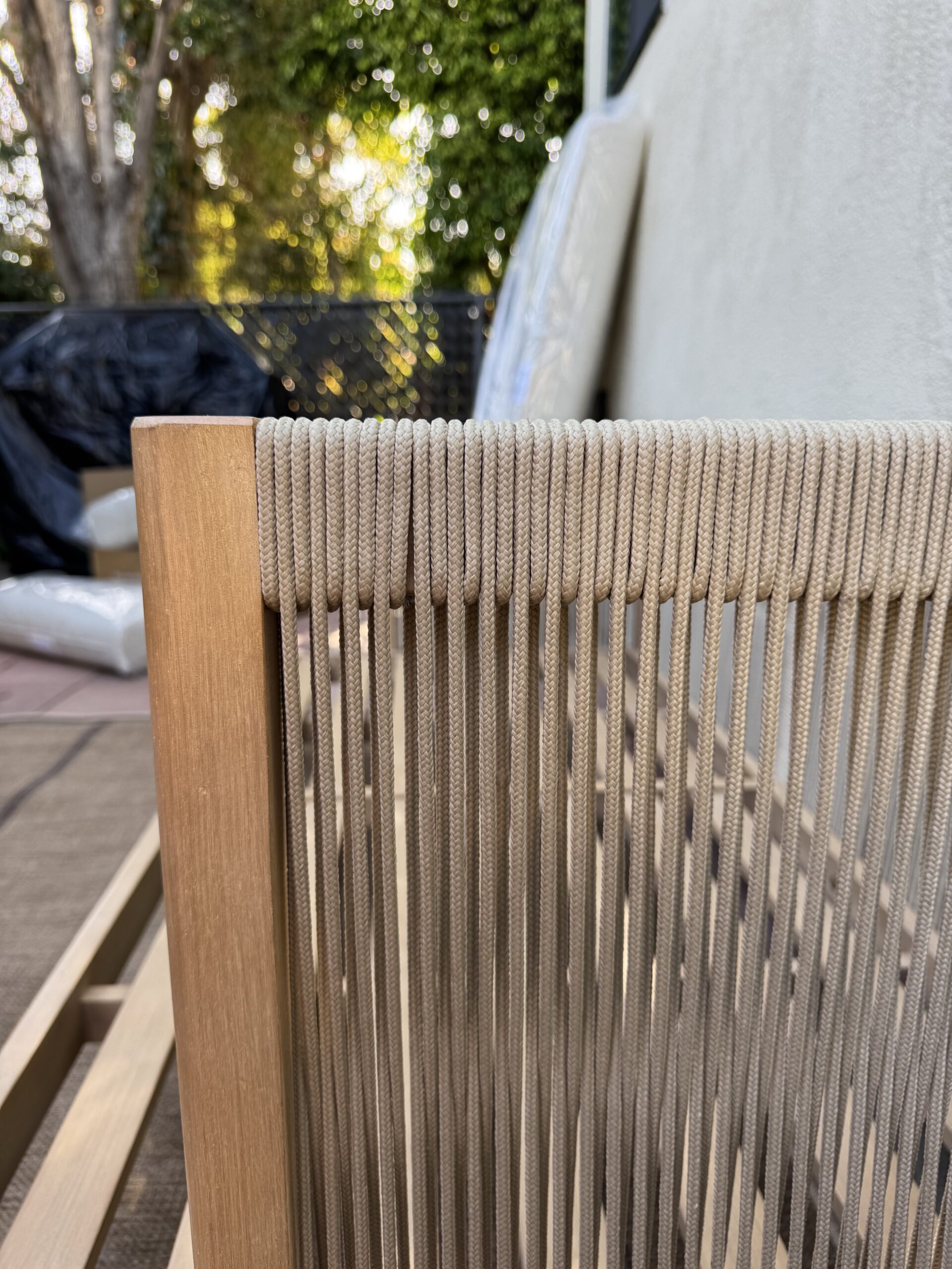 Close-up of a wooden chair or bench with tightly woven beige cord on the armrest, set outdoors near a white wall and greenery.