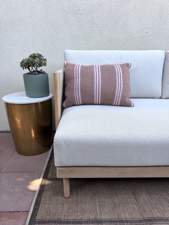 A modern outdoor sofa with a white cushion and a striped pillow sits next to a gold side table with a potted succulent plant.