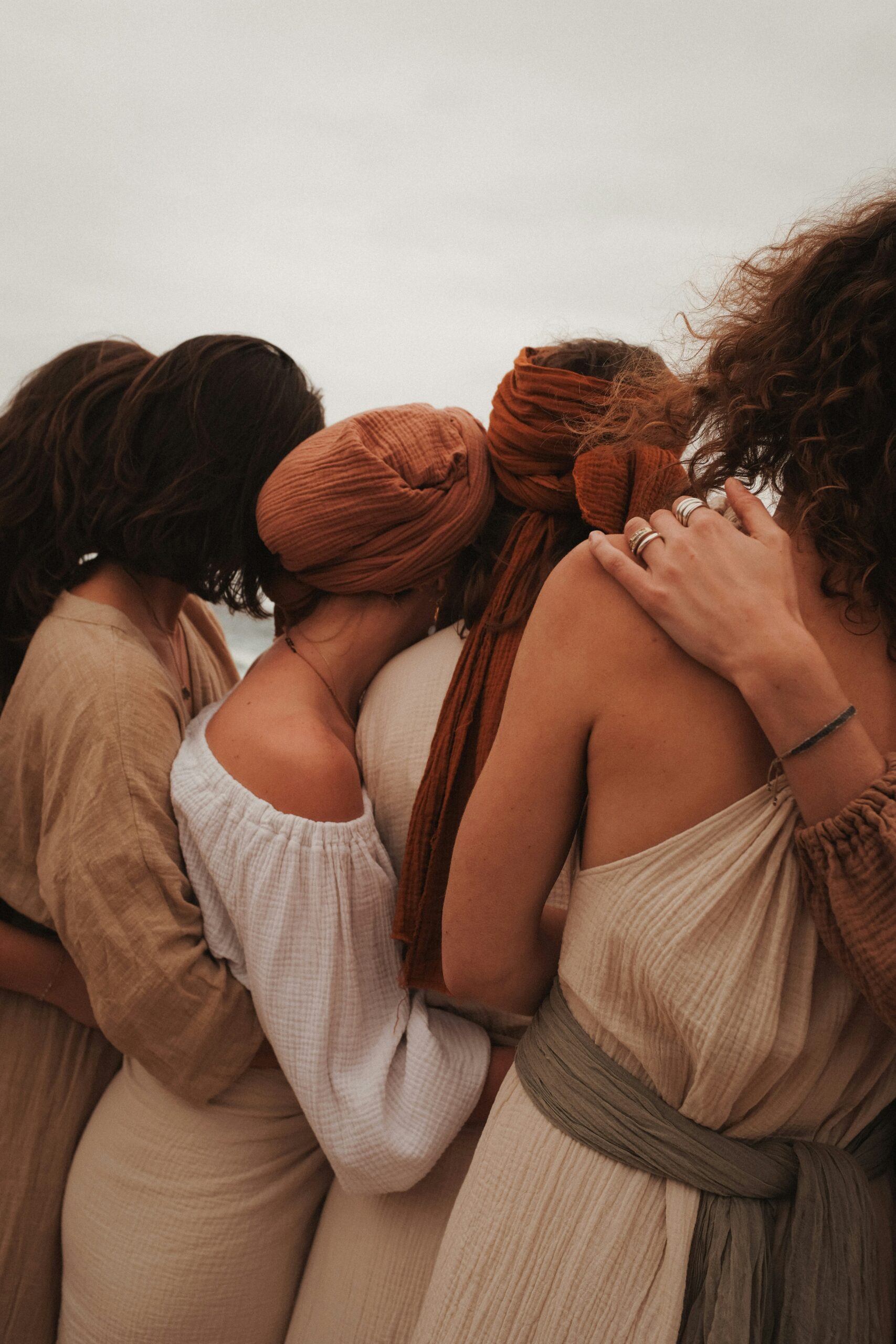 Four women dressed in neutral-toned clothing stand closely together, facing away from the camera with arms around each other.