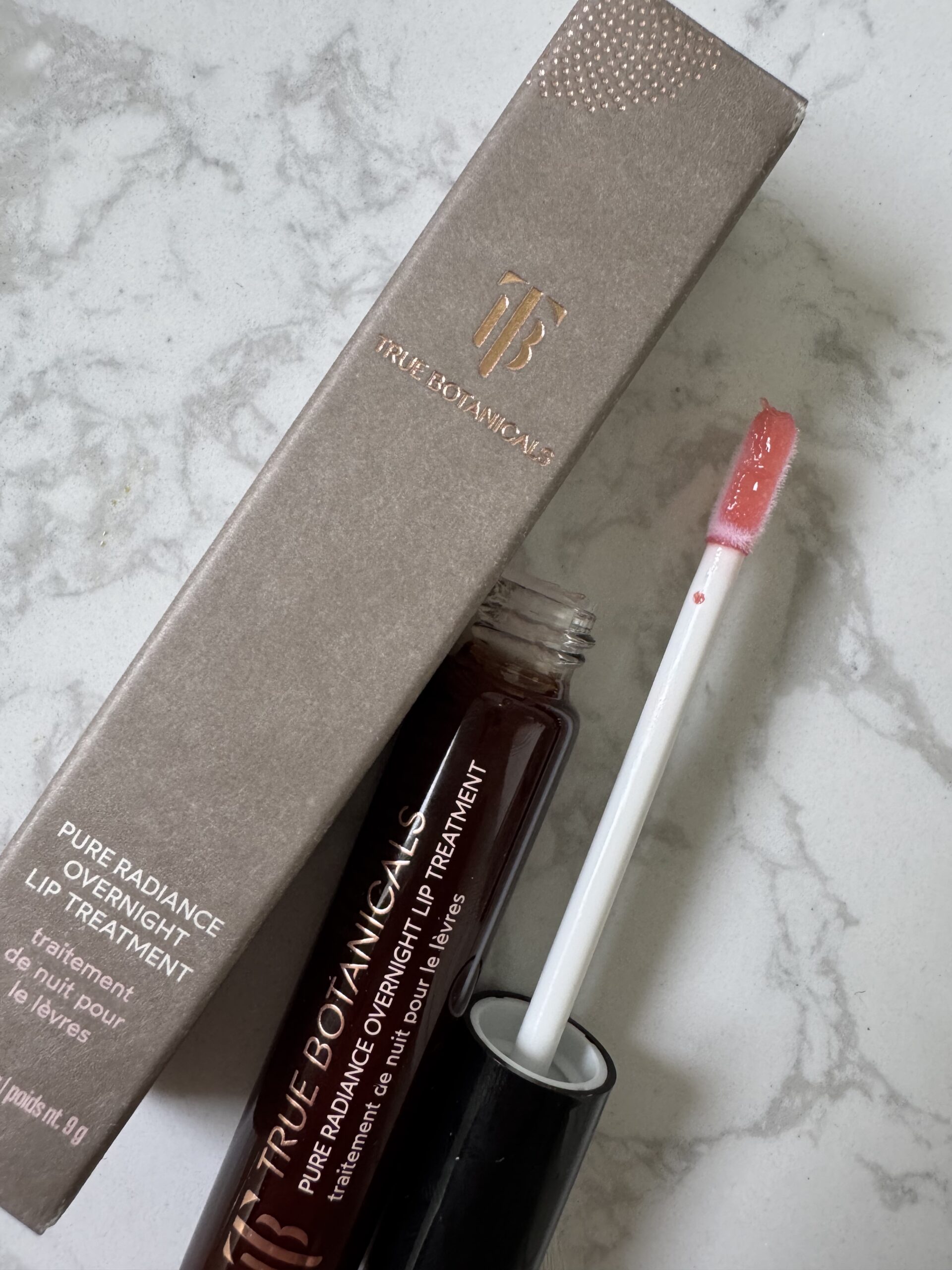 An open tube of True Botanicals Pure Radiance Overnight Lip Treatment with its applicator, next to the product's packaging, on a marble surface.