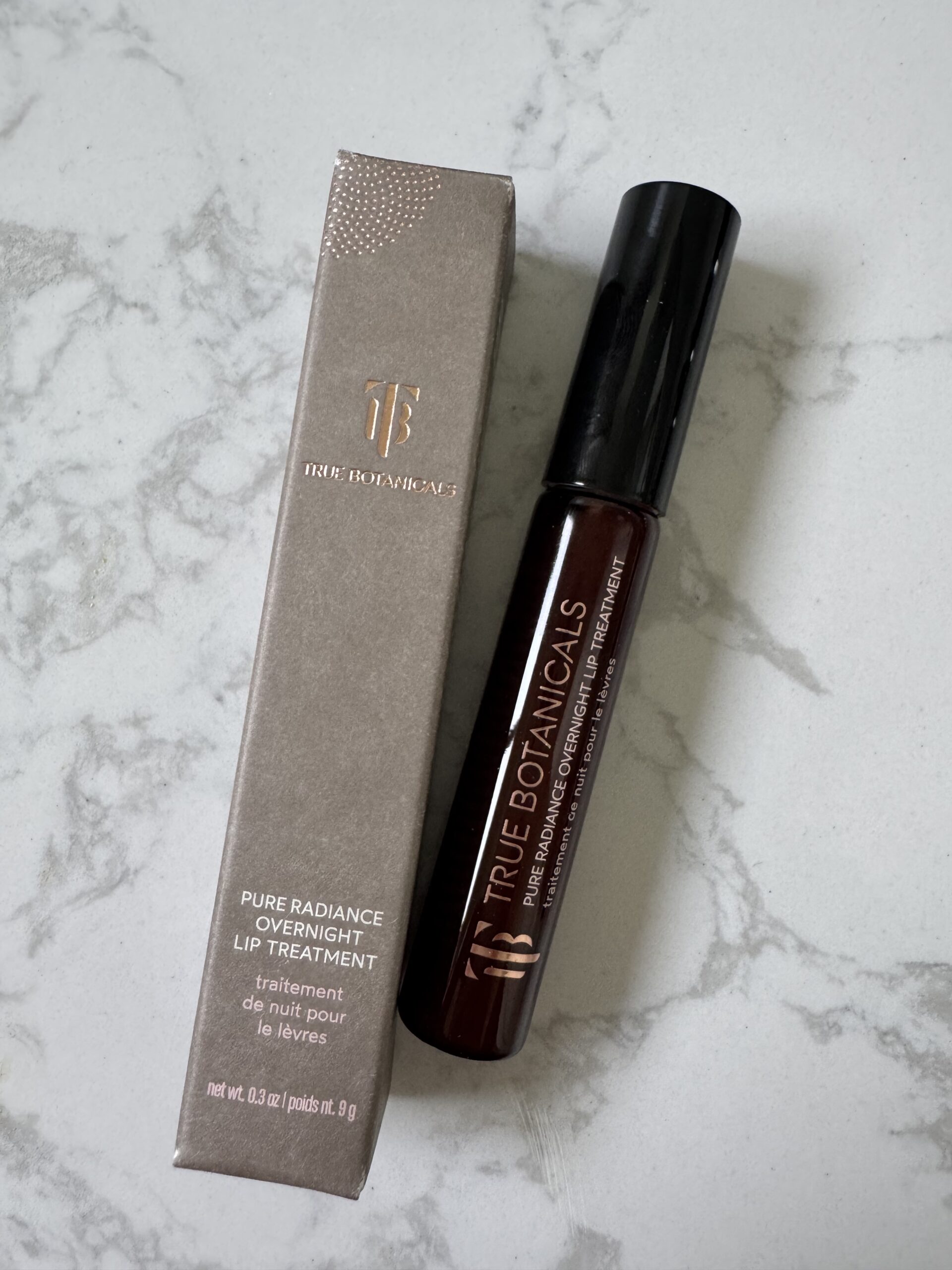 A True Botanicals Pure Radiance Overnight Lip Treatment tube is placed next to its packaging on a white marble surface.
