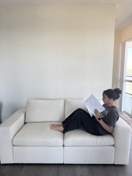 Person sitting on a white couch near a blank wall, reading a document with a window and partial outdoor view in the background.