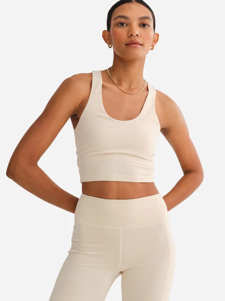 A woman wearing a beige tank top and matching high-waisted leggings stands with her hands on her hips against a plain background.