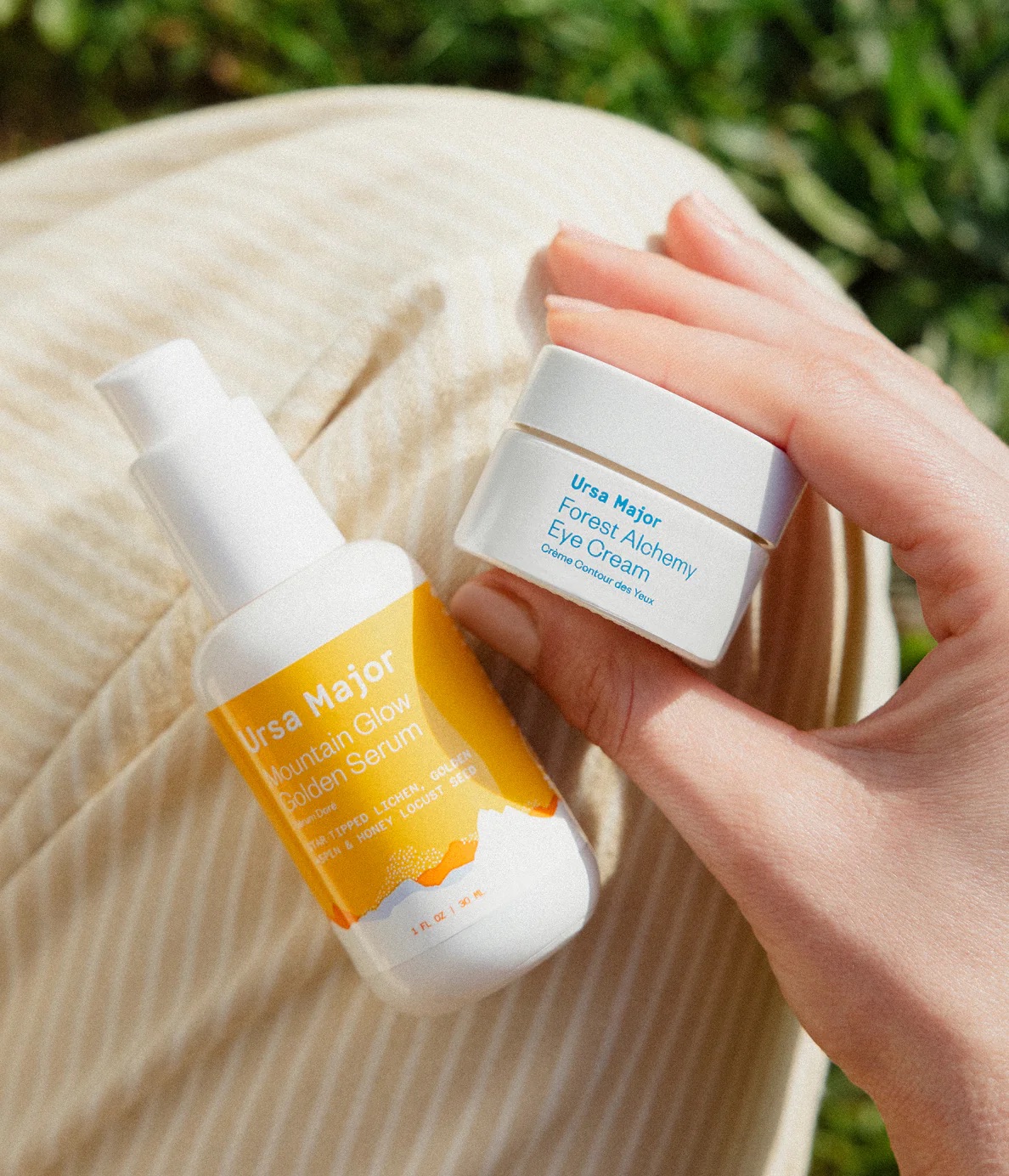 A hand holds an Ursa Major Forest Alchemy Eye Cream jar next to a bottle of Ursa Major Mountain Glow Golden Serum, resting on pinstriped pants outdoors.