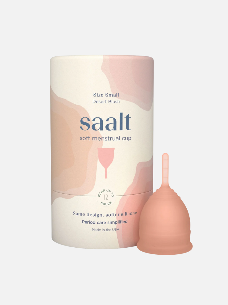 A Saalt soft menstrual cup in small size is shown next to its cylindrical packaging, which features soft pink and beige abstract designs and product information.