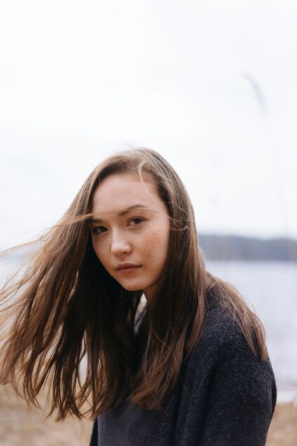 A young woman with long brown hair looks at the camera outdoors on a cloudy day, with wind blowing her hair and a blurred natural background.