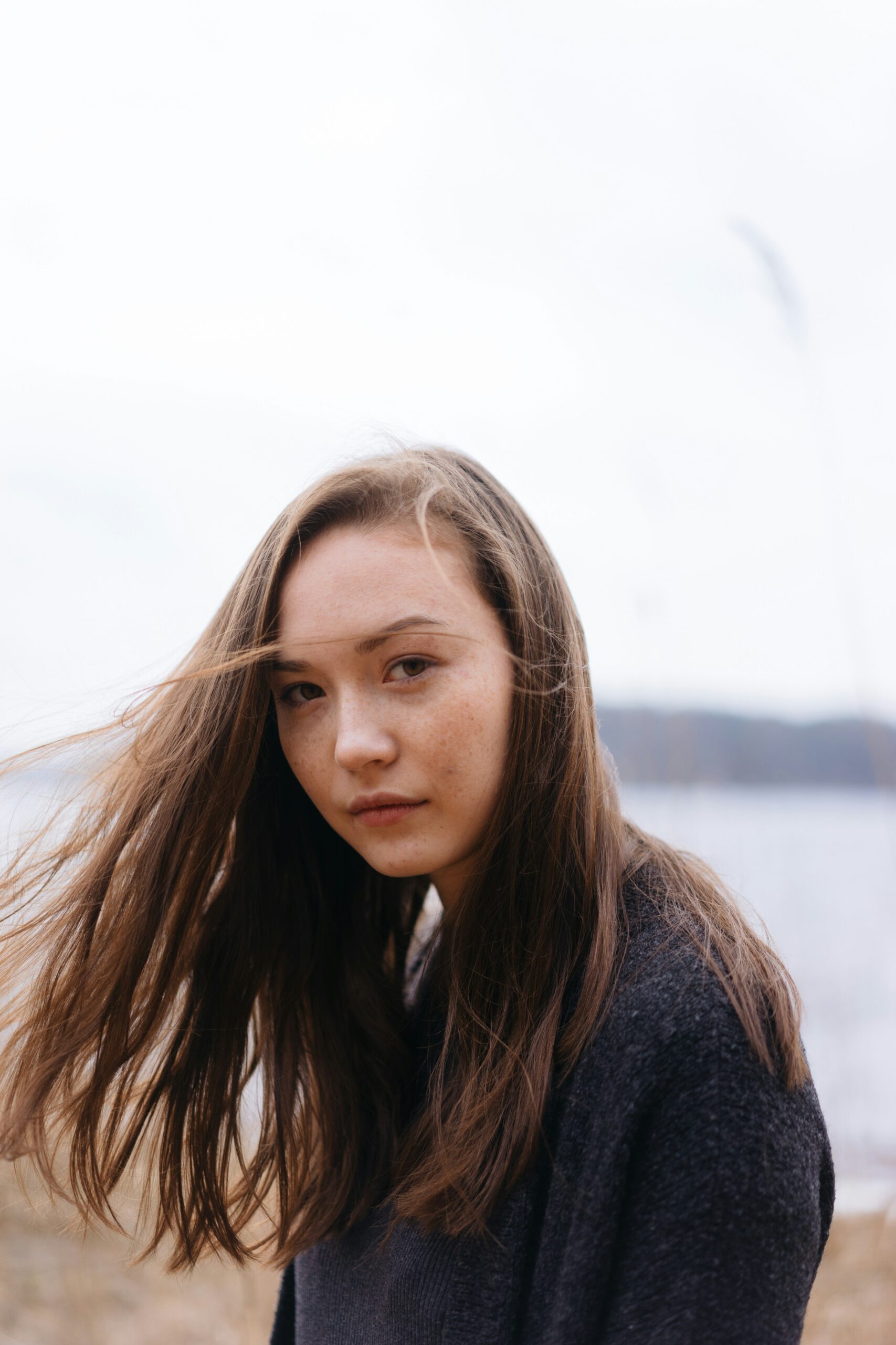 A young woman with long brown hair looks at the camera outdoors on a cloudy day, with wind blowing her hair and a blurred natural background.
