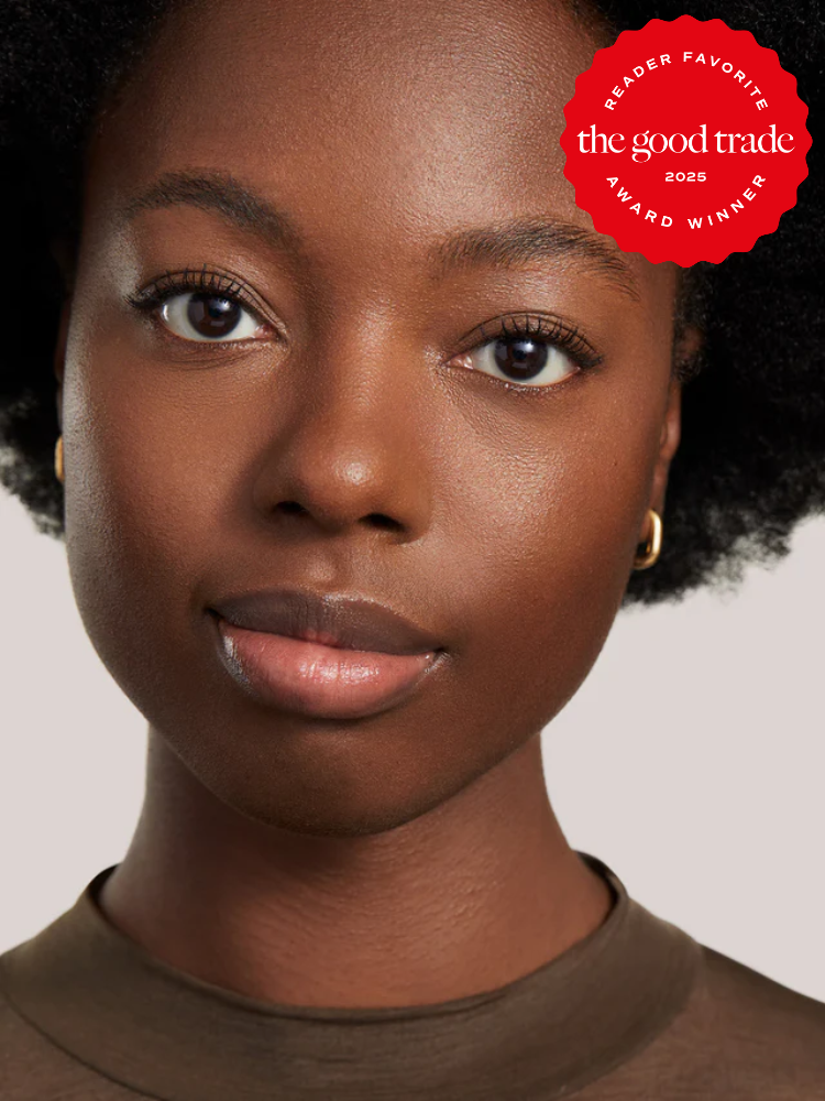 A person with dark skin and short curly hair looks at the camera, wearing a brown top and gold earrings. A red "The Good Trade Award Winner 2025" badge appears in the corner.
