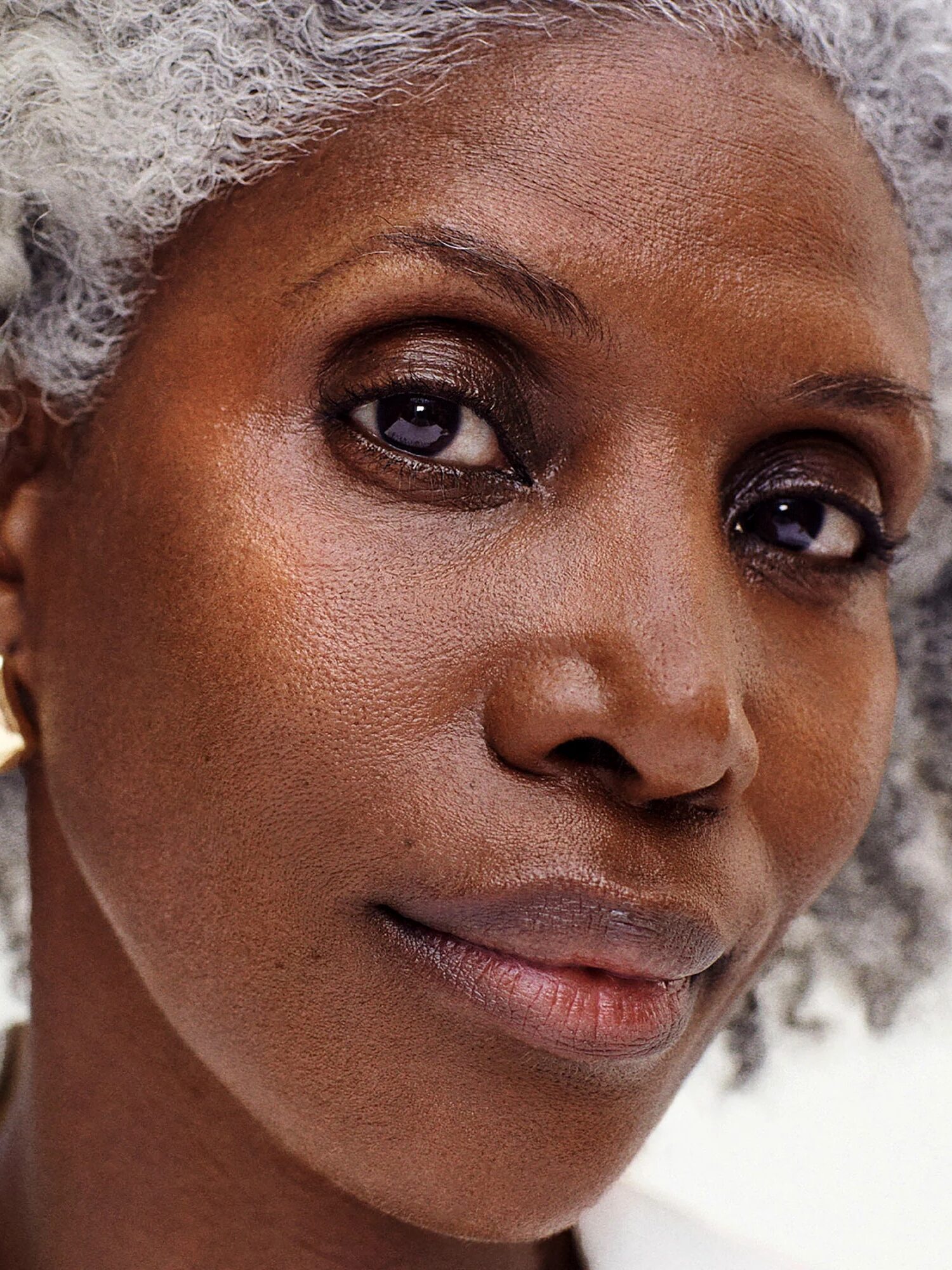 Close-up of an older woman with gray curly hair, brown skin, gold hoop earrings, and a neutral expression, looking directly at the camera.