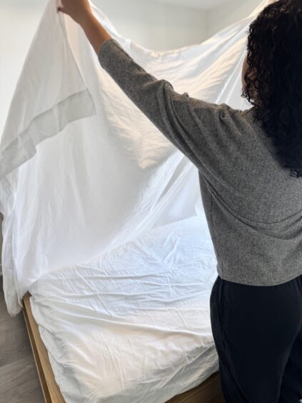 A person is spreading a white sheet over a bed, preparing to make the bed in a bright, tidy room.