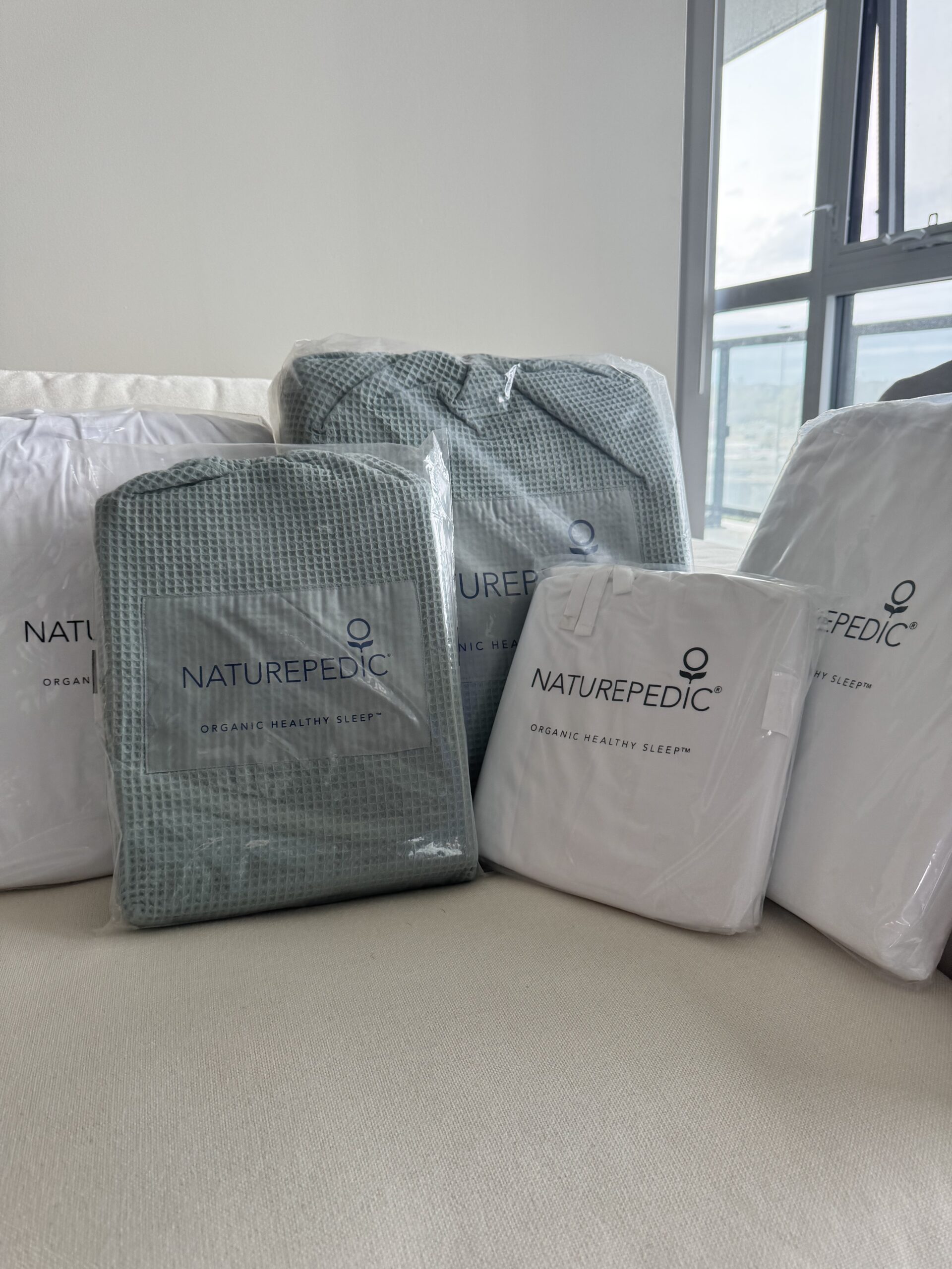 Several packaged Naturepedic bedding items, including white and green sets, are arranged on a beige couch near a window in a bright room.