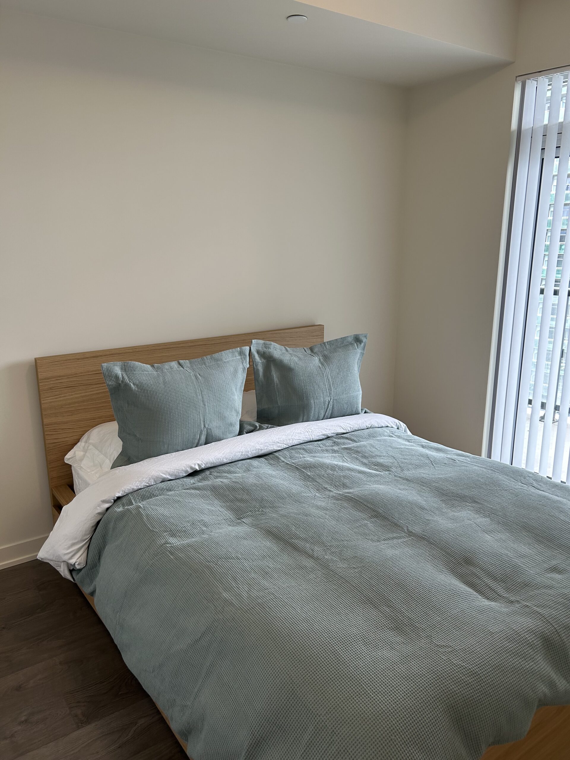 A neatly made bed with green bedding and two pillows in a minimally decorated room with light walls and a window with vertical blinds.