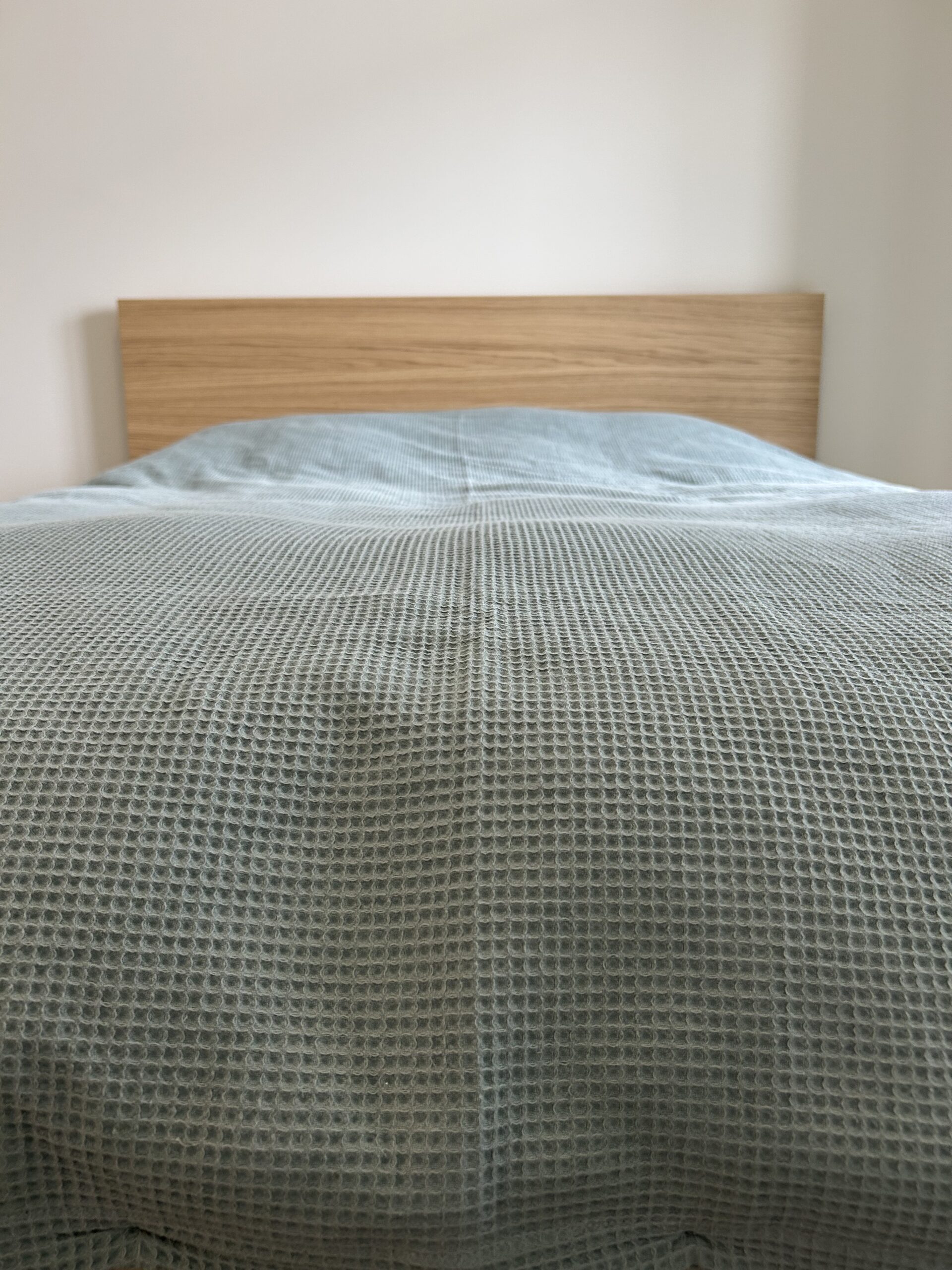 A neatly made bed with a wooden headboard and a textured light blue bedspread, shown from the foot of the bed.