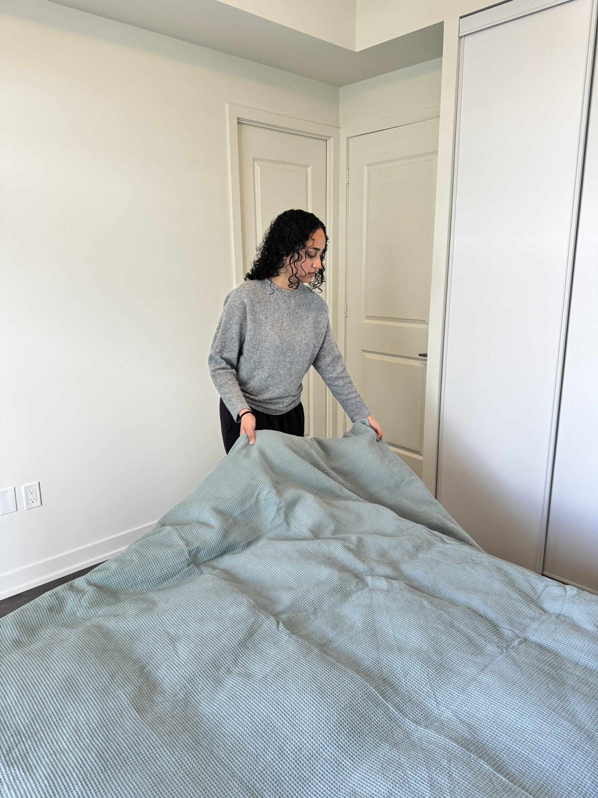 A person in a gray sweater is making a bed, straightening a blue blanket in a minimalist bedroom with white walls and doors.
