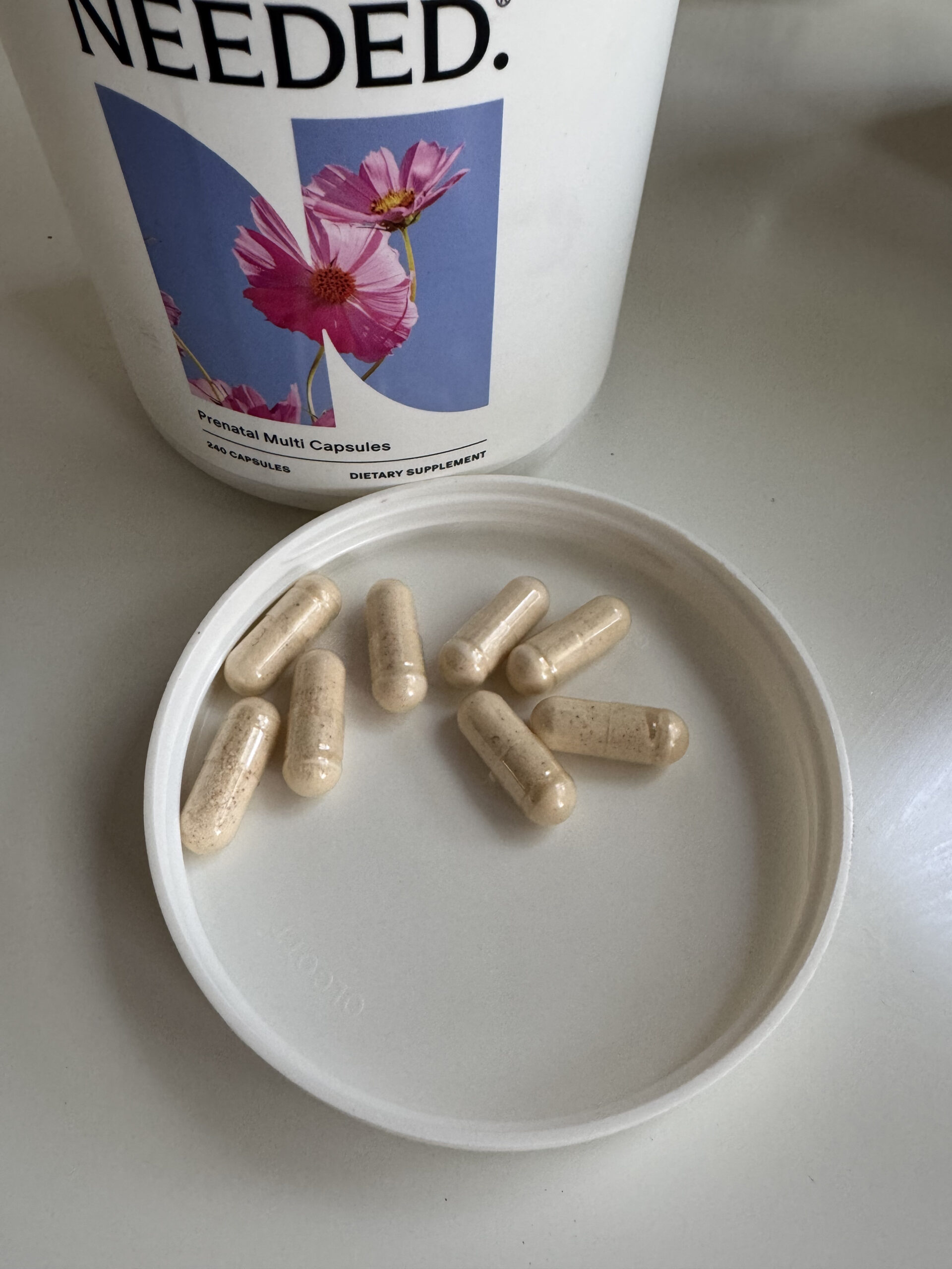 Eight beige capsules rest on a white lid next to a container labeled "Needed. Prenatal Multi Capsules," a prenatal vitamin with a floral design.