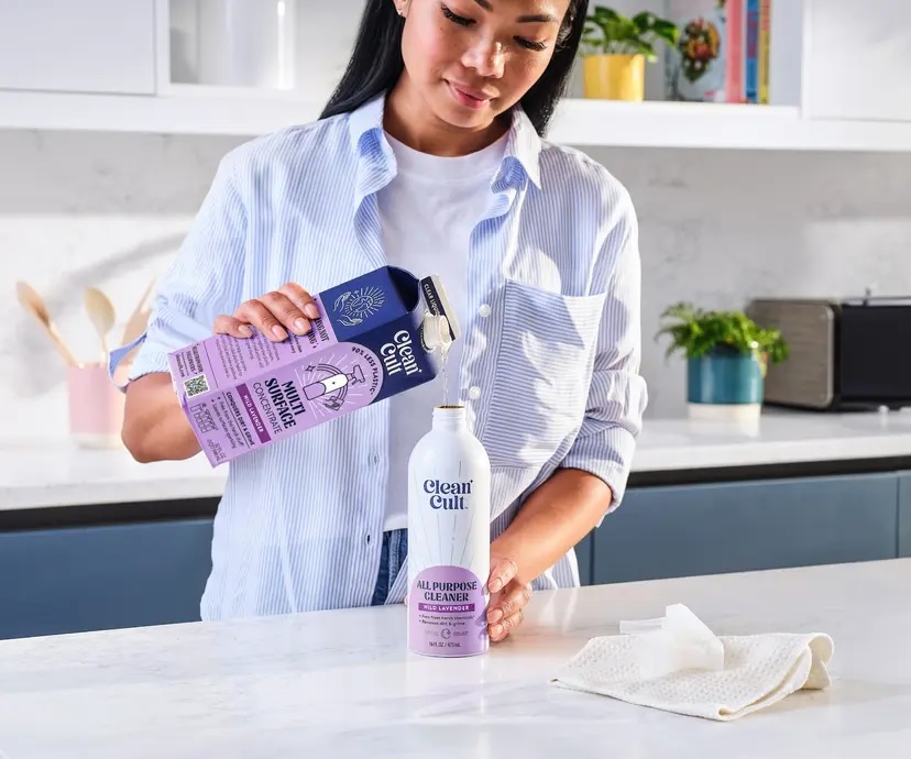 A woman pours Cleancult all-purpose cleaner from a refill carton into a bottle in a modern kitchen. A cloth is on the white countertop nearby.