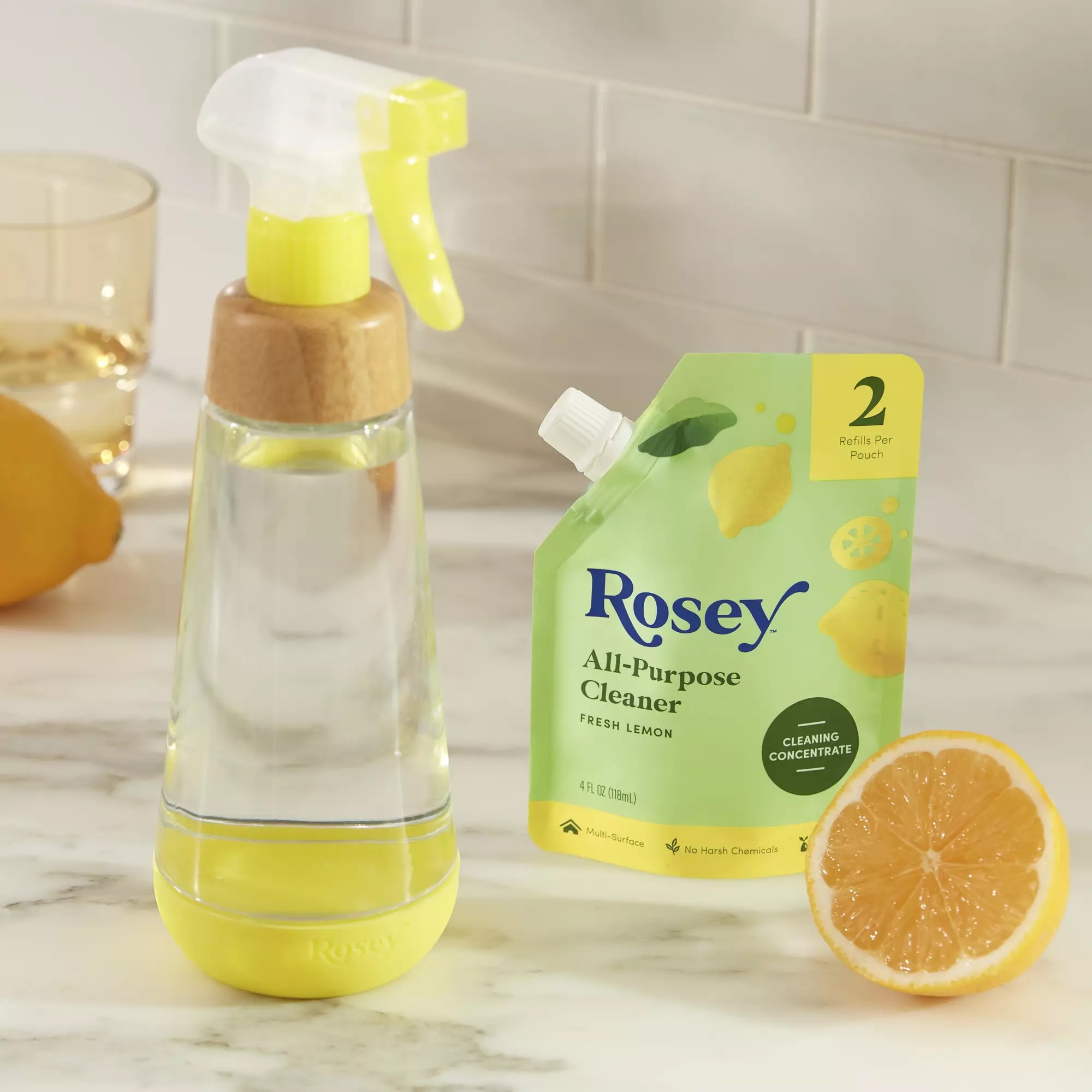 A spray bottle, a pouch of Rosey All-Purpose Cleaner, a sliced lemon, and a glass of liquid are arranged on a marble countertop.