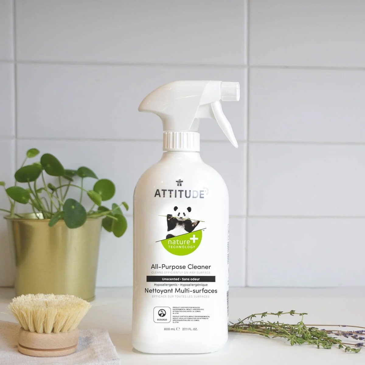 A bottle of Attitude All-Purpose Cleaner sits on a kitchen counter next to a scrub brush and a small potted plant, with a white tiled backsplash in the background.