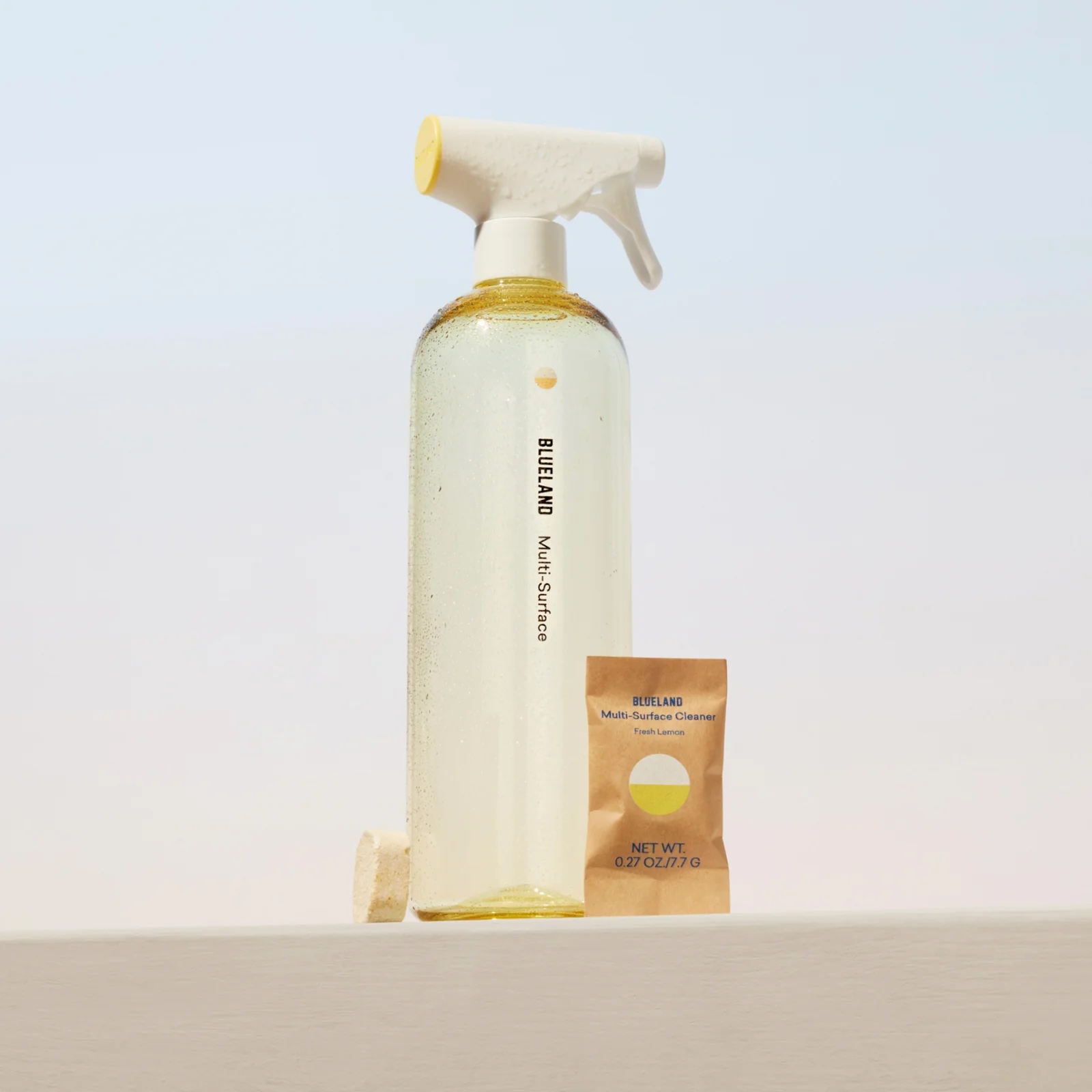 A clear spray bottle labeled "Blueland Multi-Surface" is filled with liquid, next to a packet labeled "Blueland Multi-Surface Cleaner" and a small tablet, all on a white surface.