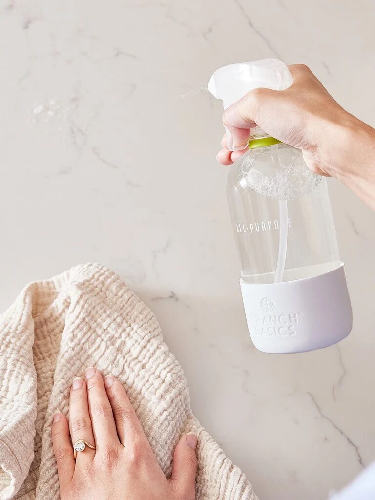 A hand sprays cleaner from a clear bottle onto a marble countertop while another hand wipes the surface with a textured cloth.