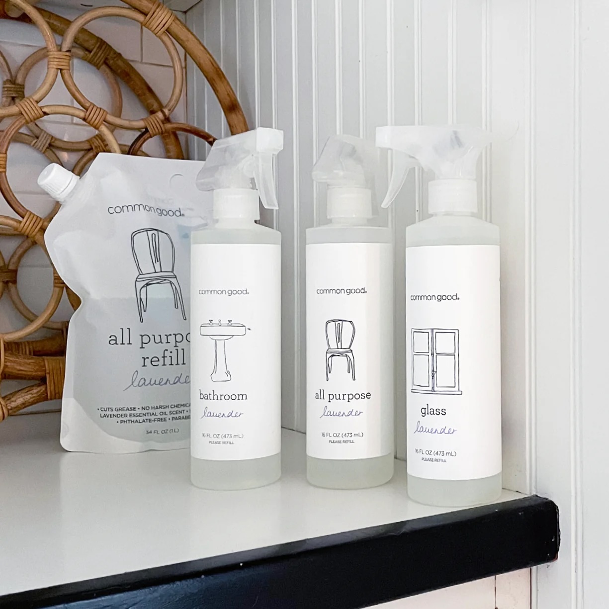 Four white bottles of Common Good cleaning products, including an all purpose refill pouch, are displayed on a white shelf against a white paneled wall with a wicker decor piece.
