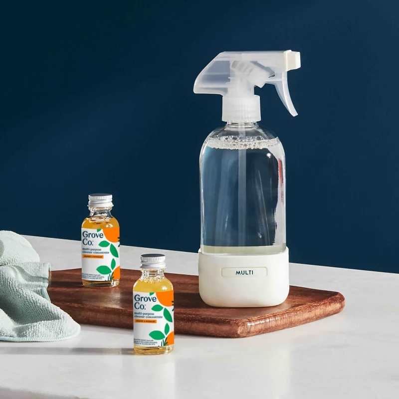 A spray bottle labeled "MULTI" stands on a wooden tray next to two Grove Co. cleaning solution bottles and a folded cloth on a white surface against a dark blue background.