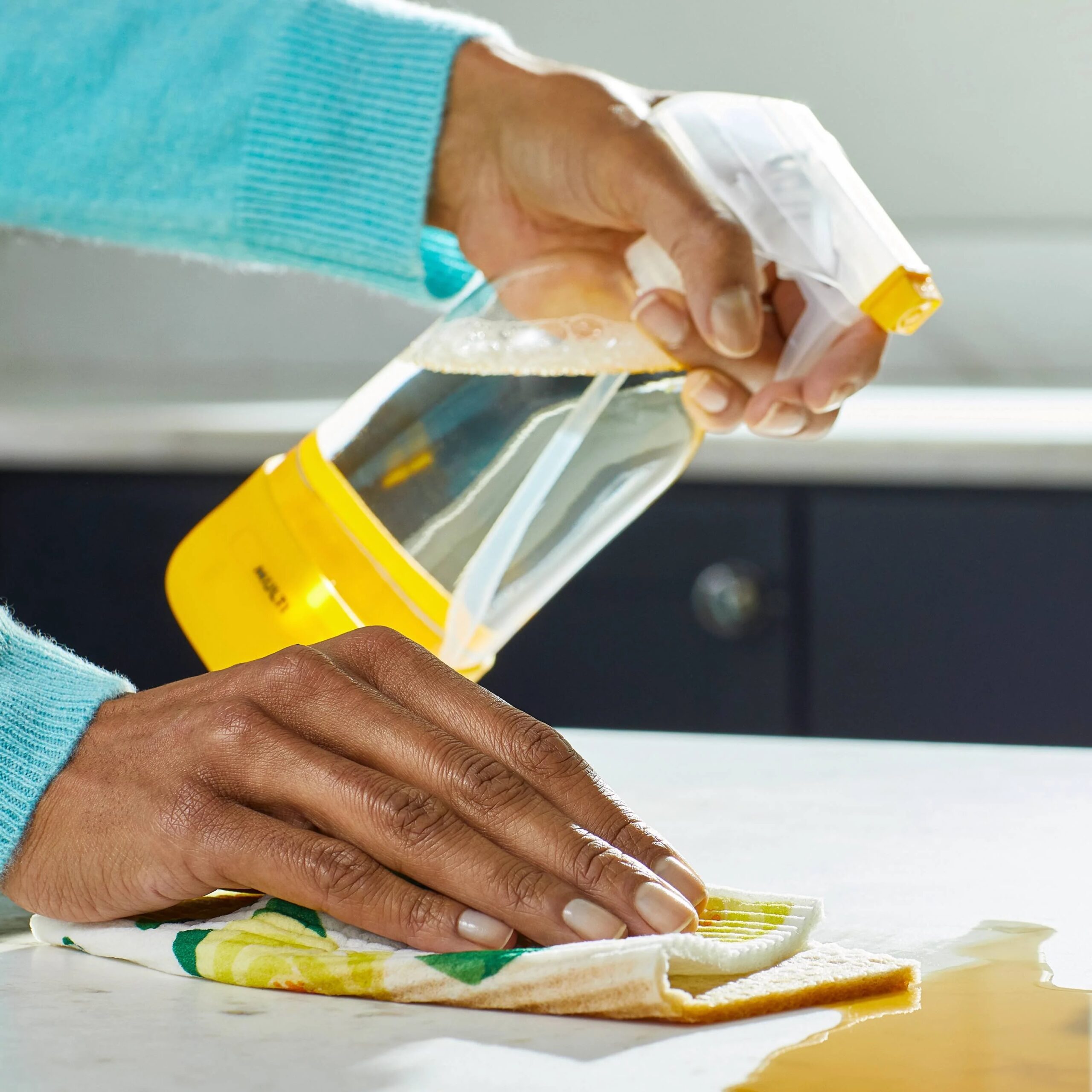 A person cleaning a countertop with a yellow spray bottle and a cloth, wiping up a spill.