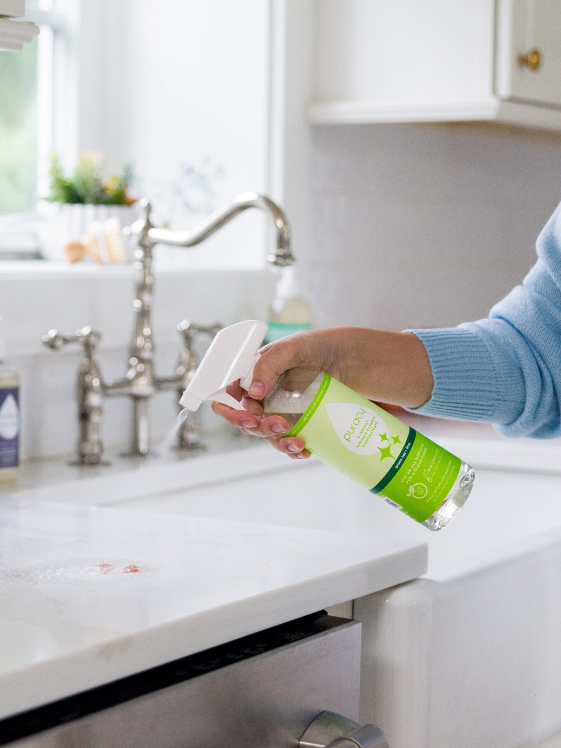 A person sprays a green-labeled cleaner onto a white kitchen countertop near a gray cloth, with a sink and soap dispenser in the background.