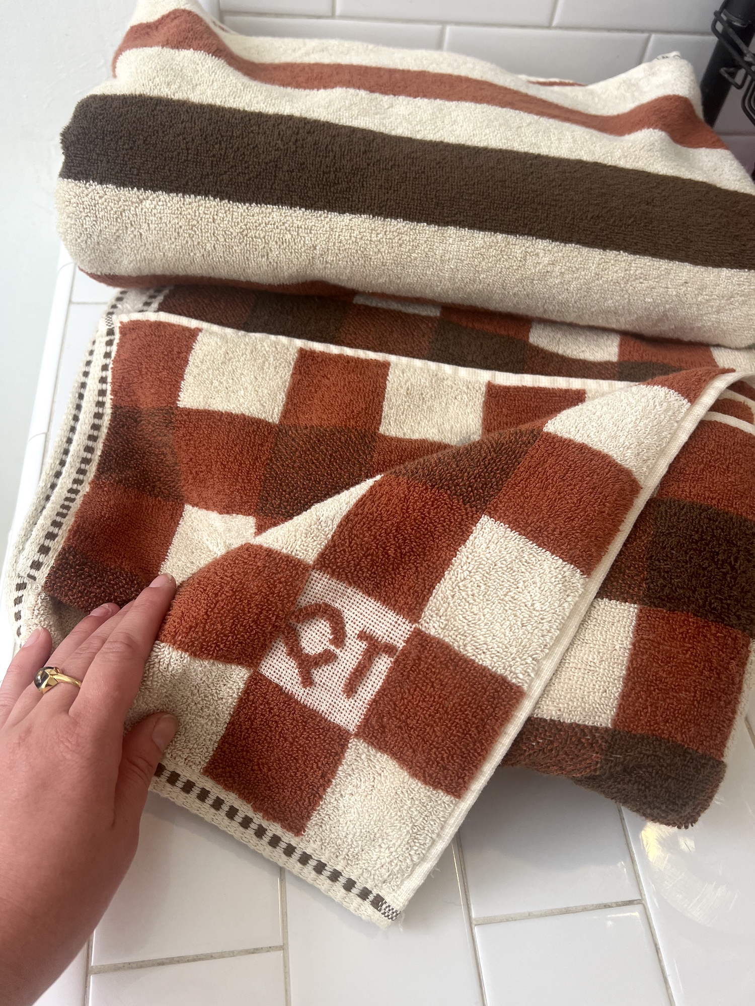 A hand touches a folded checkered organic towel with "QT" woven into it, next to a striped towel, both on a white tiled surface.