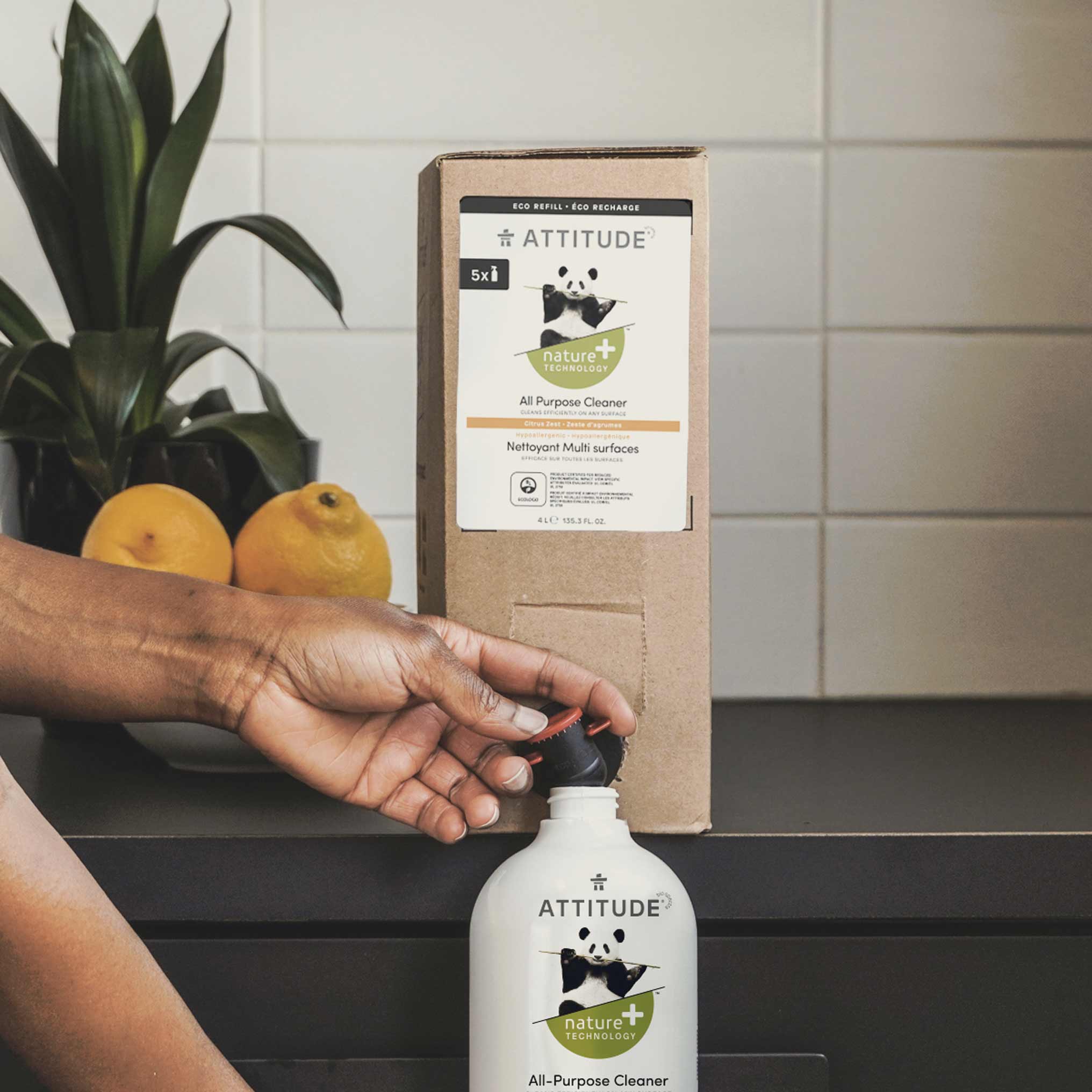 A person refills an Attitude Nature+ All-Purpose Cleaner bottle from a larger box, with lemons and a tiled wall in the background.