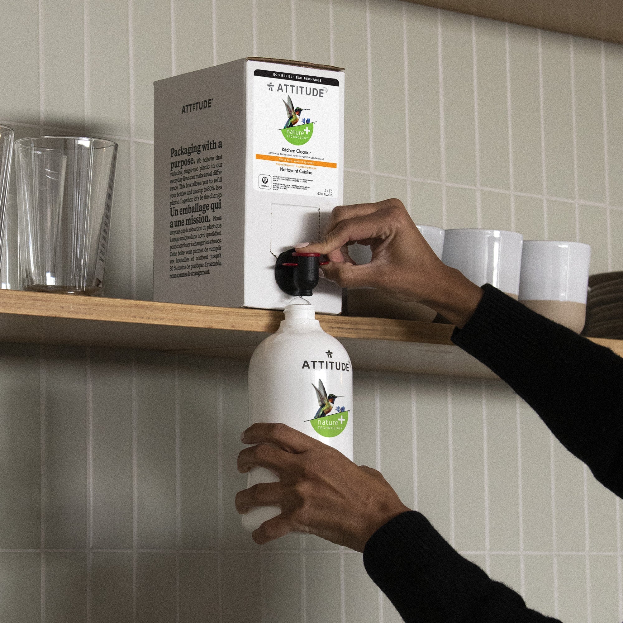 A person dispenses liquid from an "ATTITUDE" box container into a white refillable bottle in a kitchen with glasses and cups on a wooden shelf.