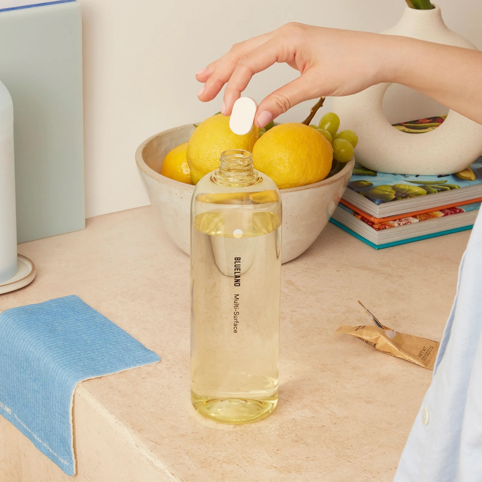 A hand drops a white tablet into a clear bottle labeled "BLUELAND Multi-Surface," with a bowl of fruit and some books in the background.