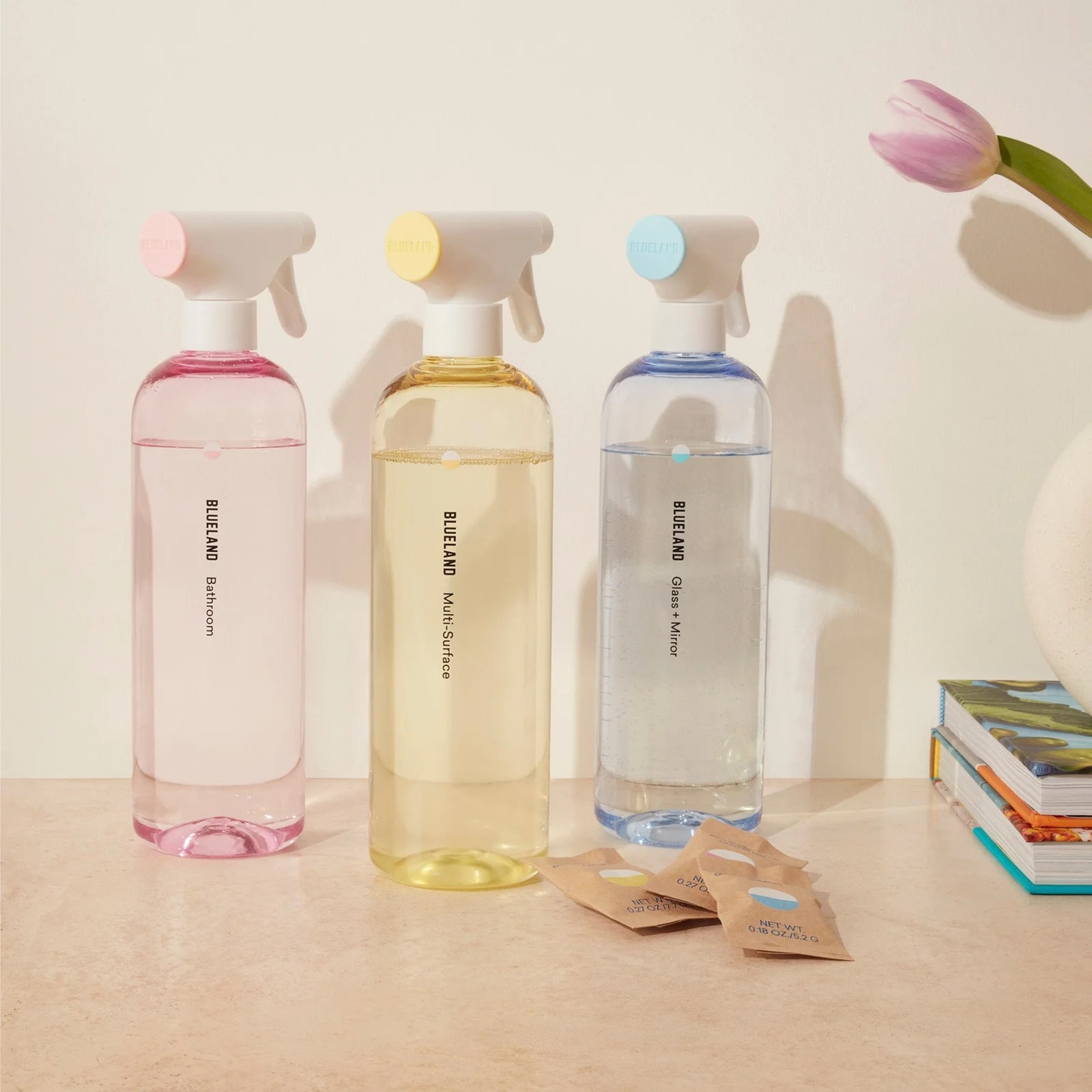 Three refillable spray bottles labeled Bathroom, Multi-Surface, and Glass & Mirror are arranged on a counter next to a flower vase, books, and cleaning refill packets.