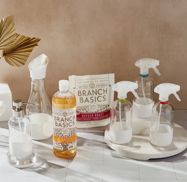 A collection of Branch Basics cleaning products, including spray bottles, a soap dispenser, a carafe, and a bottle of concentrate, arranged on a countertop with a neutral background.