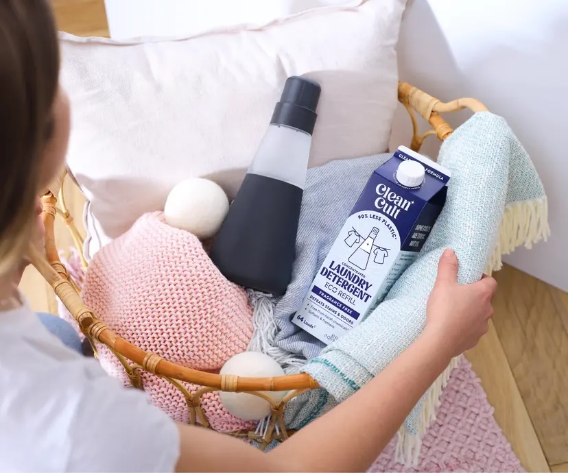 A person places a throw blanket into a laundry basket containing detergent, a bottle, dryer balls, and folded towels on a wooden floor.