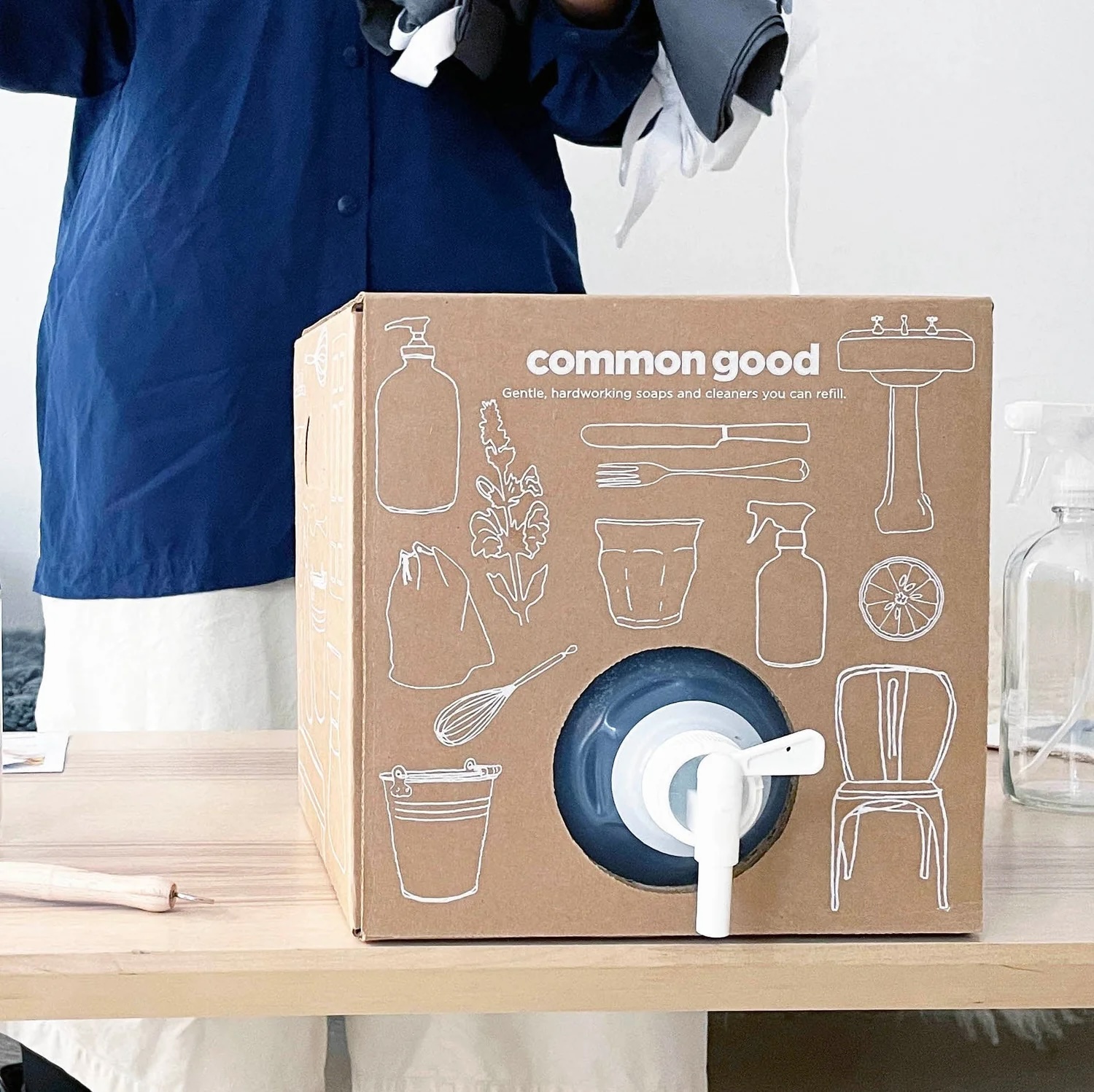 A cardboard box labeled "common good" with white line drawings of household items and a spout for dispensing liquid sits on a table; a person stands behind it.
