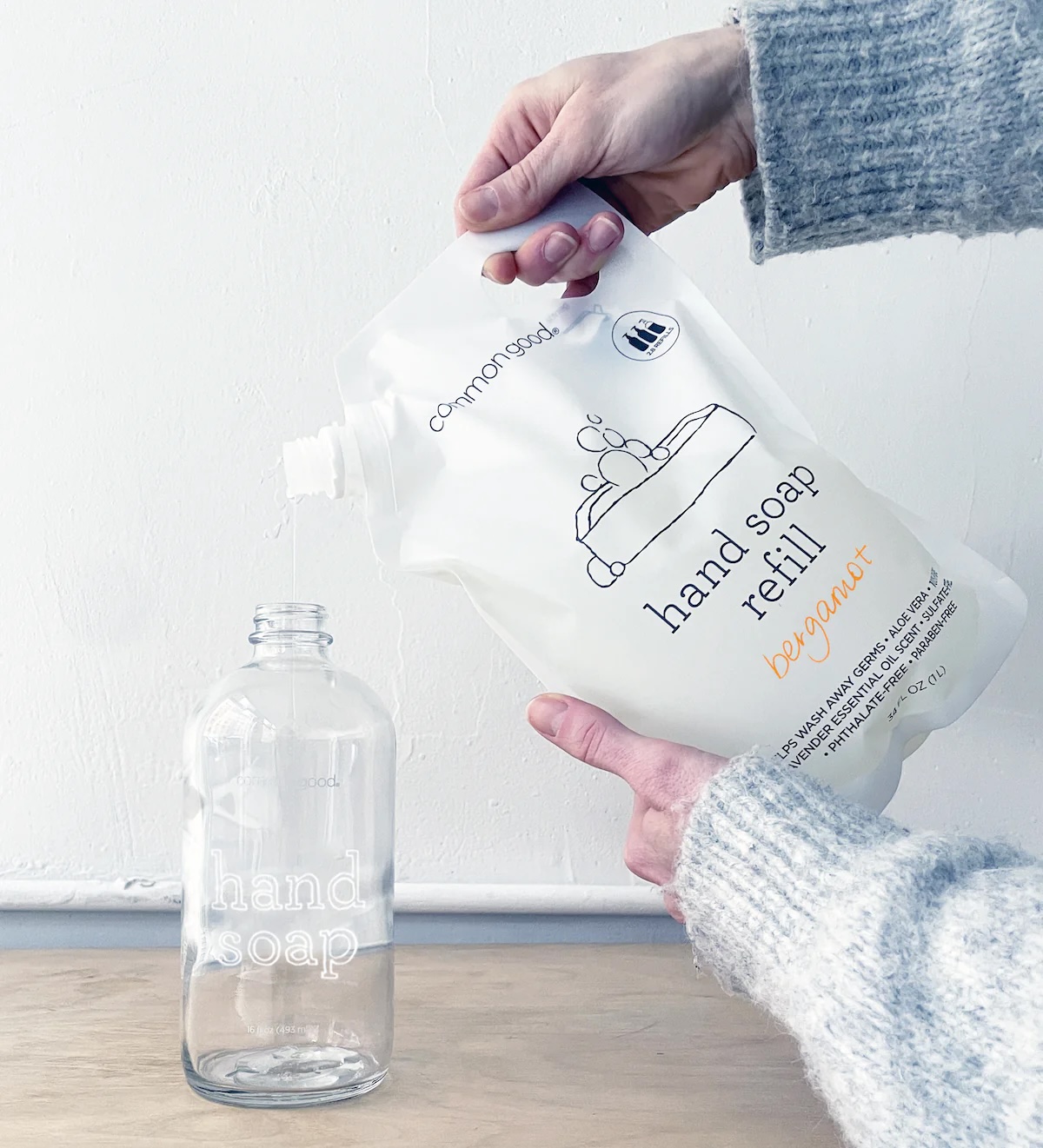 A person pours hand soap refill from a pouch labeled "hand soap refill bergamot" into an empty glass bottle labeled "hand soap.