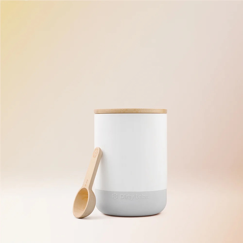 A white and gray cylindrical container with a wooden lid and a wooden measuring scoop placed beside it on a neutral background.