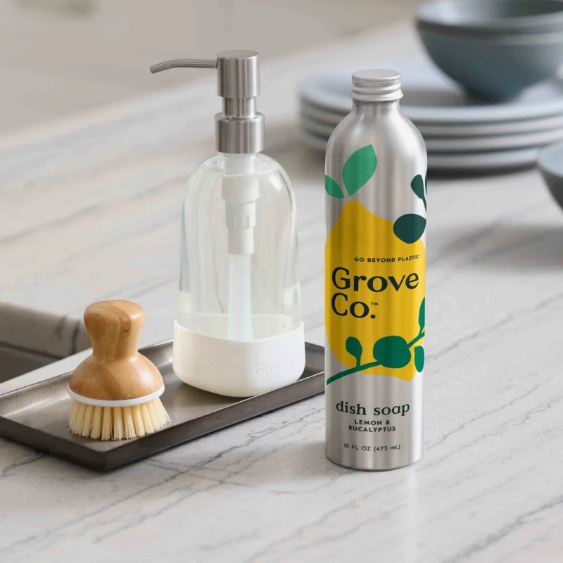 A metal bottle of Grove Co. dish soap stands next to a glass soap dispenser and a wooden dish brush on a kitchen counter.