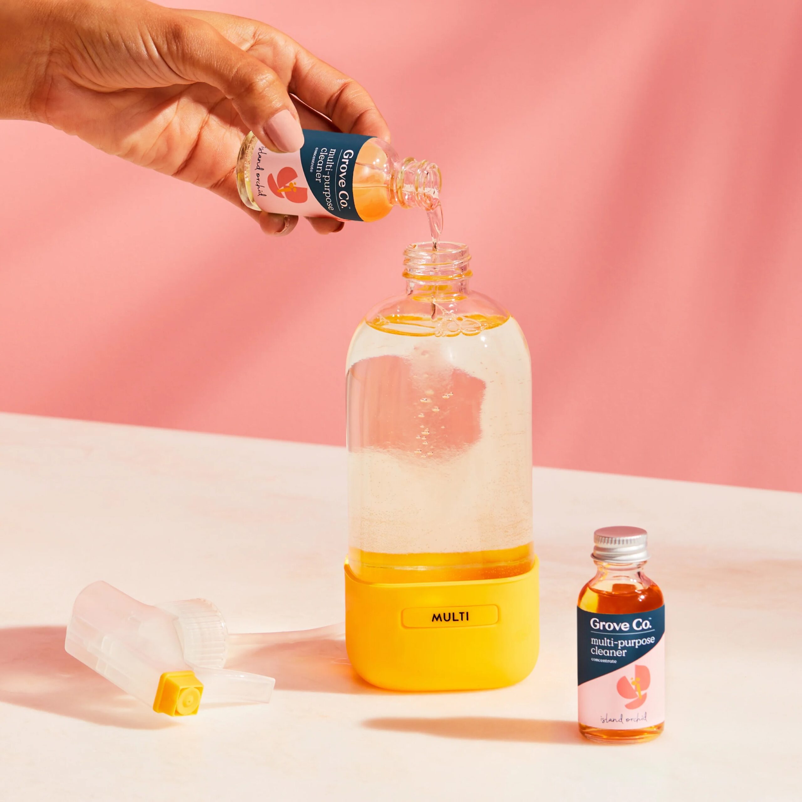 A hand pours liquid from a Grove Co. multi-purpose cleaner bottle into a clear spray bottle with a yellow base; another bottle and a nozzle lay nearby on a pink surface.