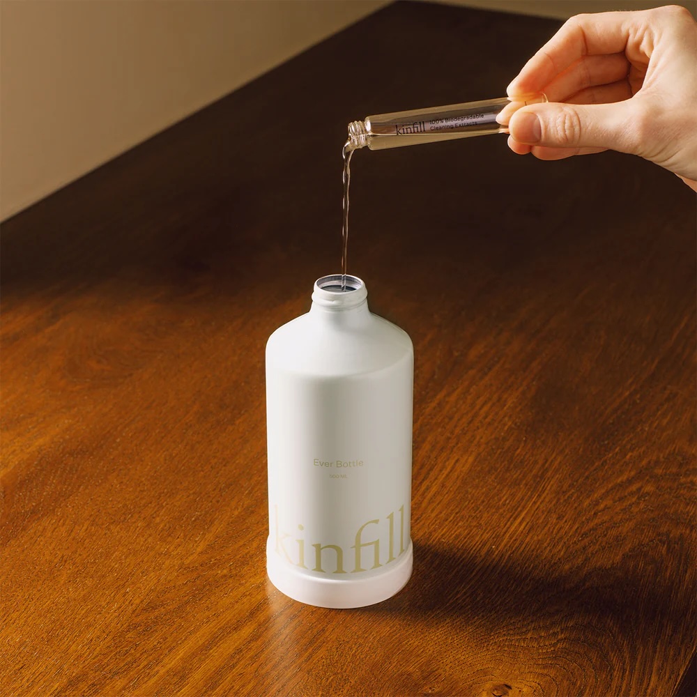 A hand pours liquid from a small vial into a white bottle labeled "skinfill" on a wooden surface.