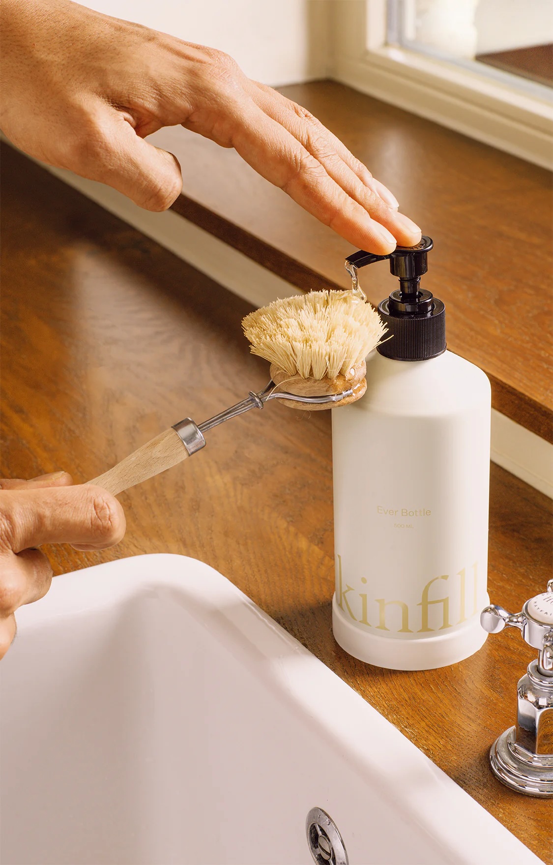 A person dispenses soap from a pump bottle labeled "skinfill" onto a dish brush over a kitchen sink.