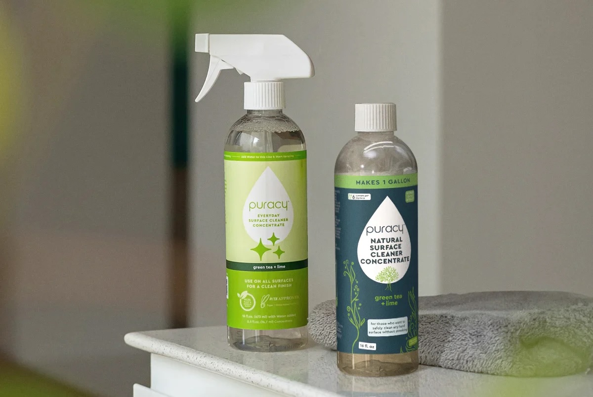 Two bottles of Puracy natural surface cleaner concentrate sit on a white countertop next to a gray towel; one bottle has a spray nozzle and the other does not.