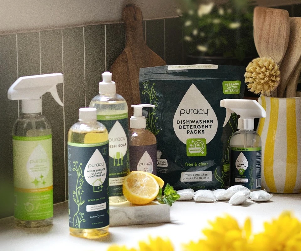 A variety of Puracy cleaning products, including sprays, soaps, and detergent packs, are arranged on a kitchen counter with a lemon, parsley, and cleaning brushes nearby.