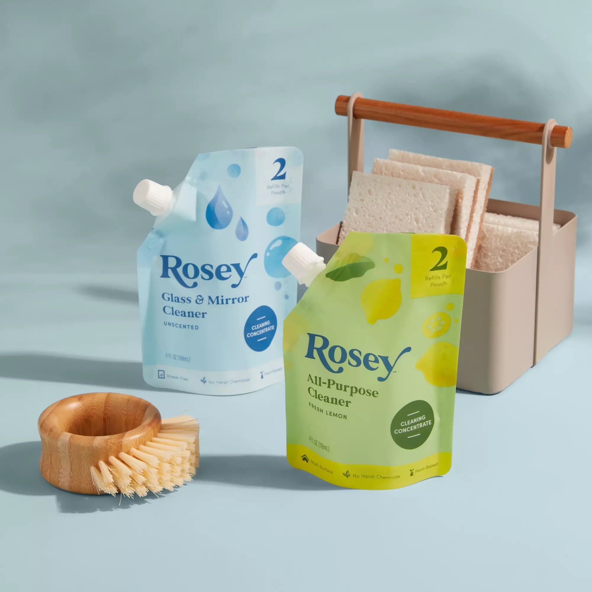 Two refill pouches of Rosey cleaners stand in front of a basket with reusable cloths and a wooden scrub brush on a light blue background.