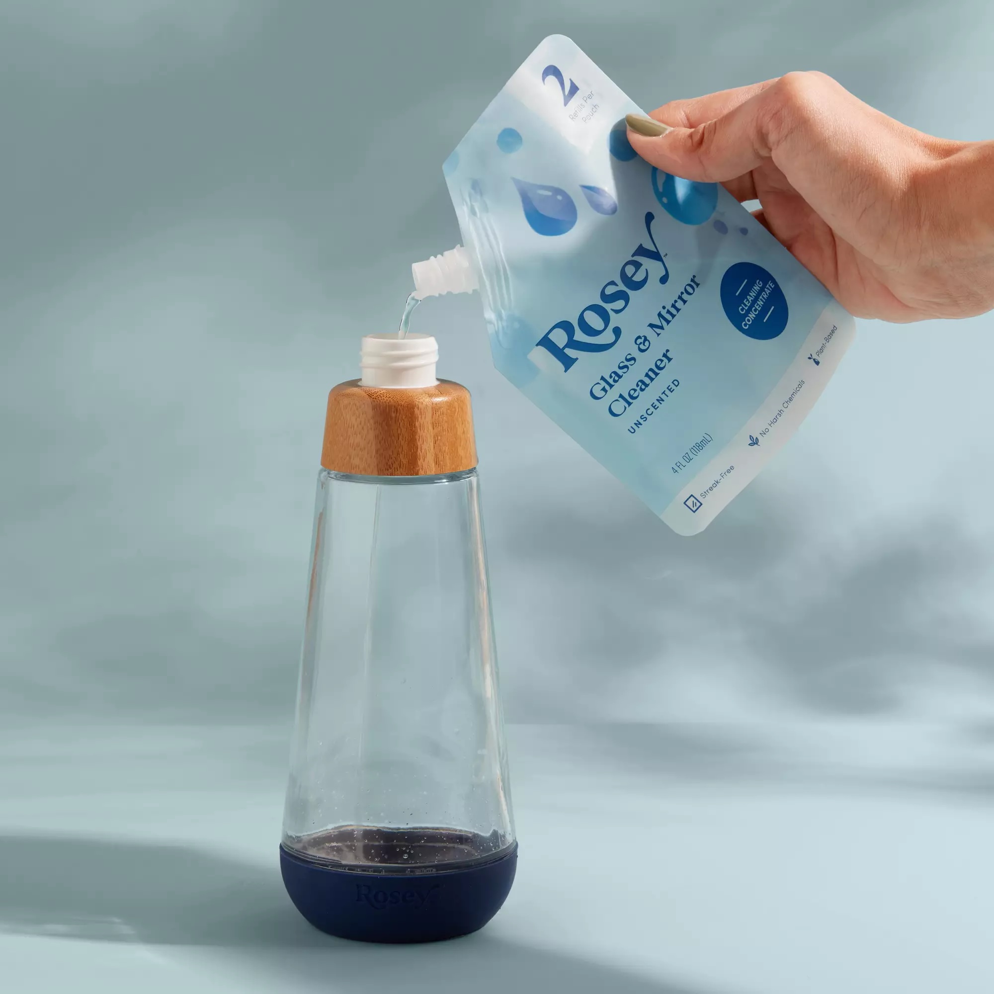 A hand pours Rosey glass and mirror cleaner from a refill pouch into a clear bottle with a wooden cap.