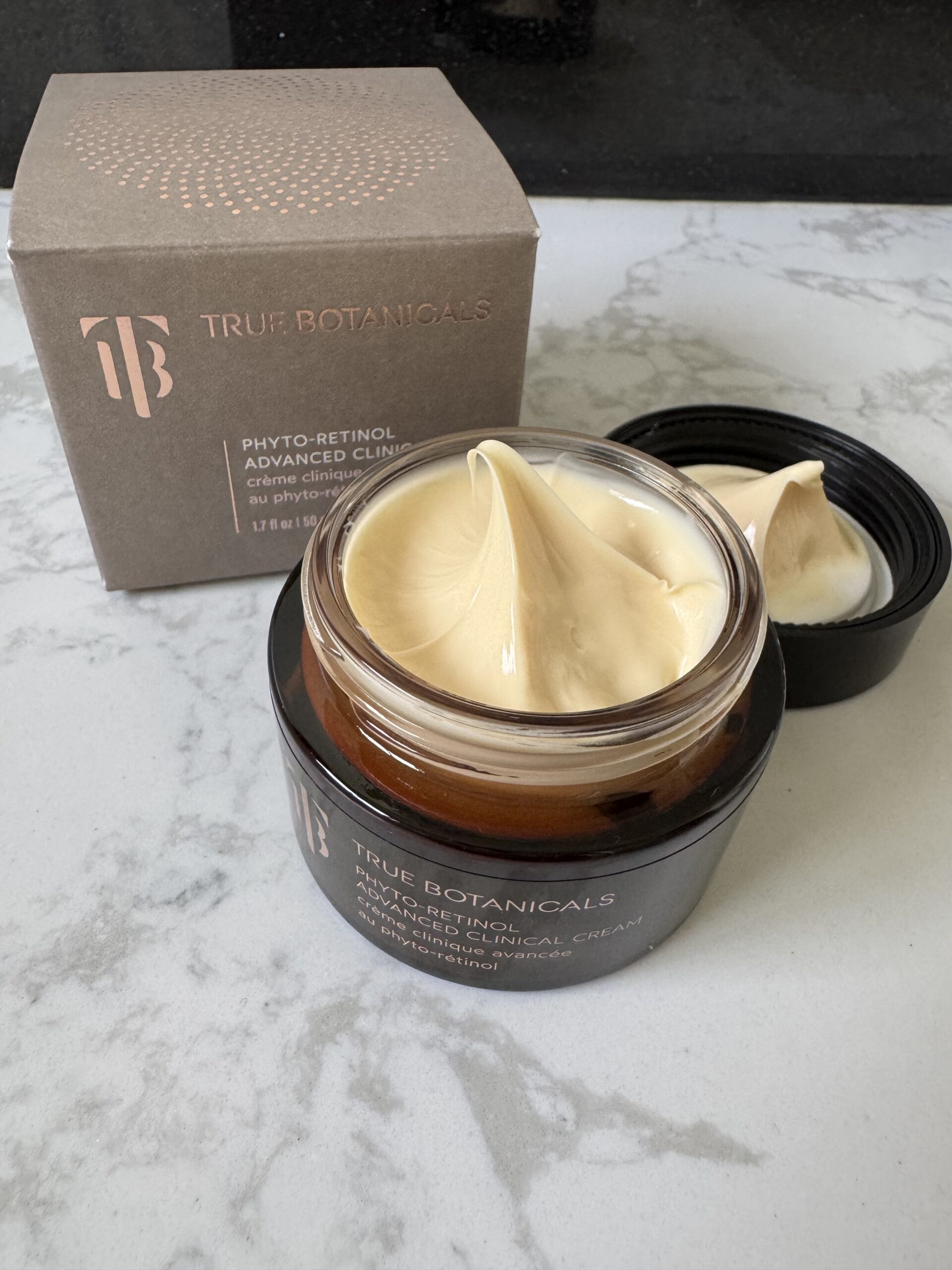 An open jar of True Botanicals Phyto-Retinol Advanced Cream sits on a marble surface next to its box, showing the smooth, creamy product inside.