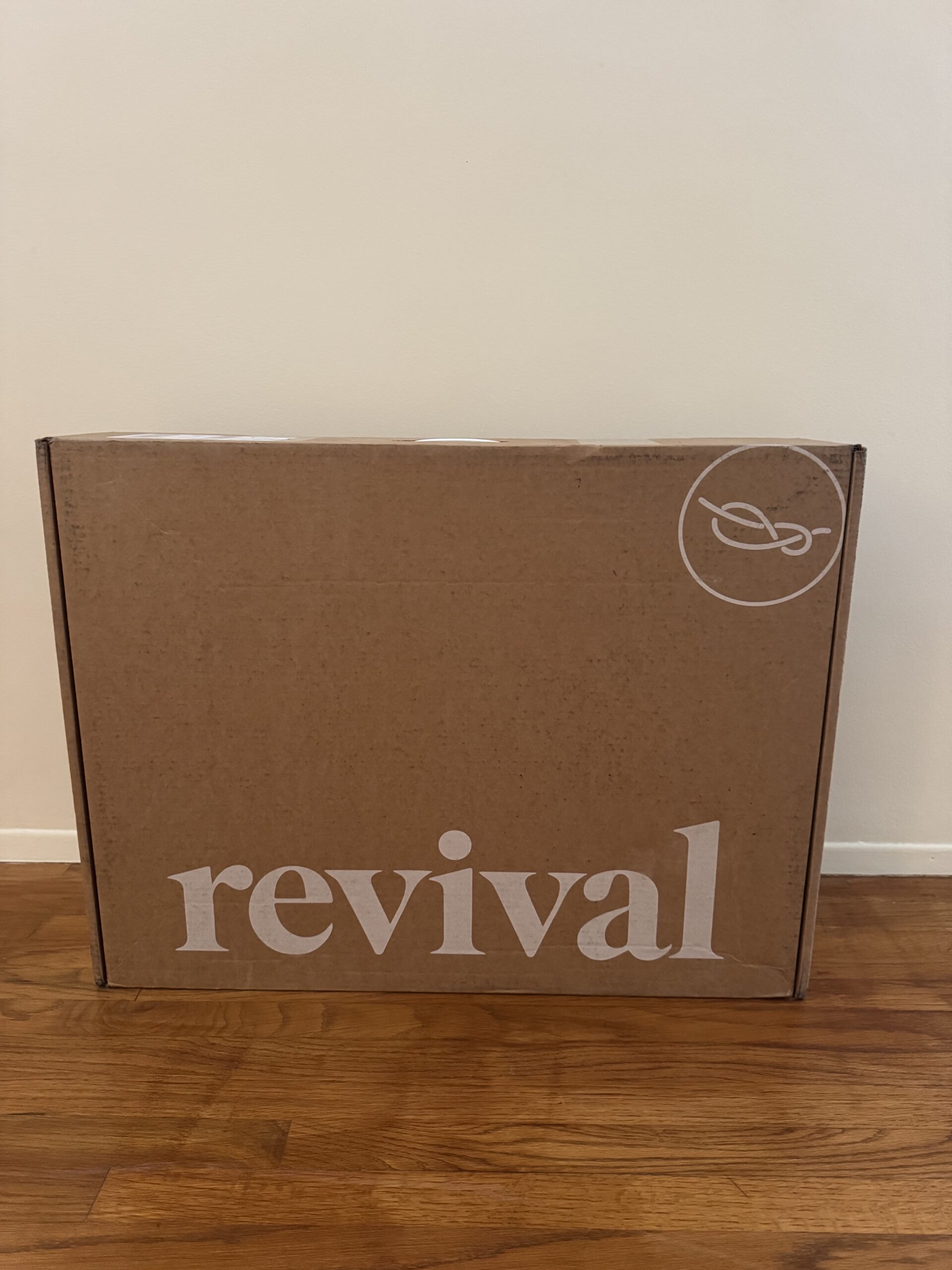A brown cardboard box with the word "revival" and a circular logo on it, placed on a wooden floor against a plain white wall.