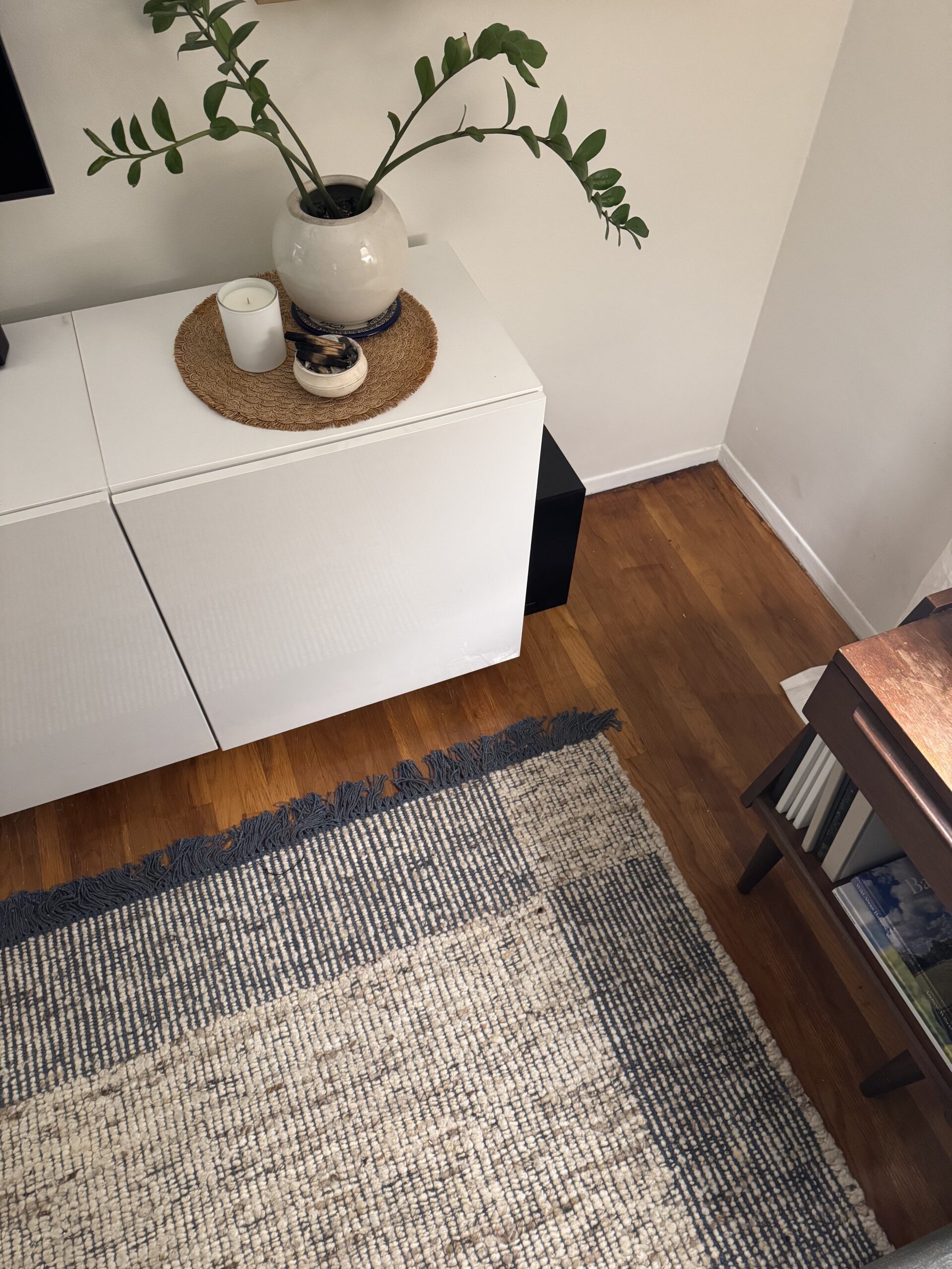 A white sideboard with a plant in a vase, a candle, and small dish sits on a hardwood floor next to a woven rug and a small bookshelf.